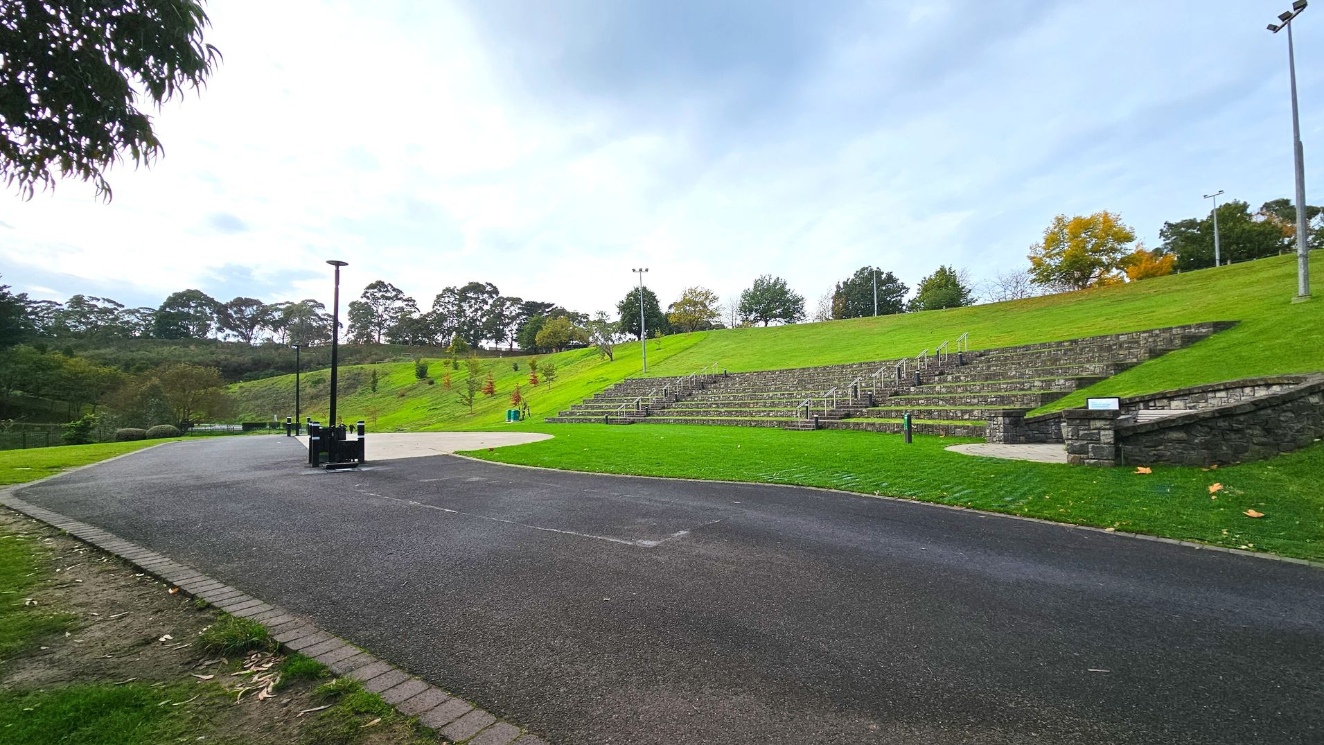 A gently sloping grassy amphitheatre with tiered stone seating on the right, surrounded by lush green grass. A wide, smooth asphalt path curves through the foreground, bordered by a narrow strip of bricks. Several tall, slender lampposts line the path, providing lighting. Trees with varying shades of green and autumnal colours are scattered across the landscape, adding depth and texture to the scene.