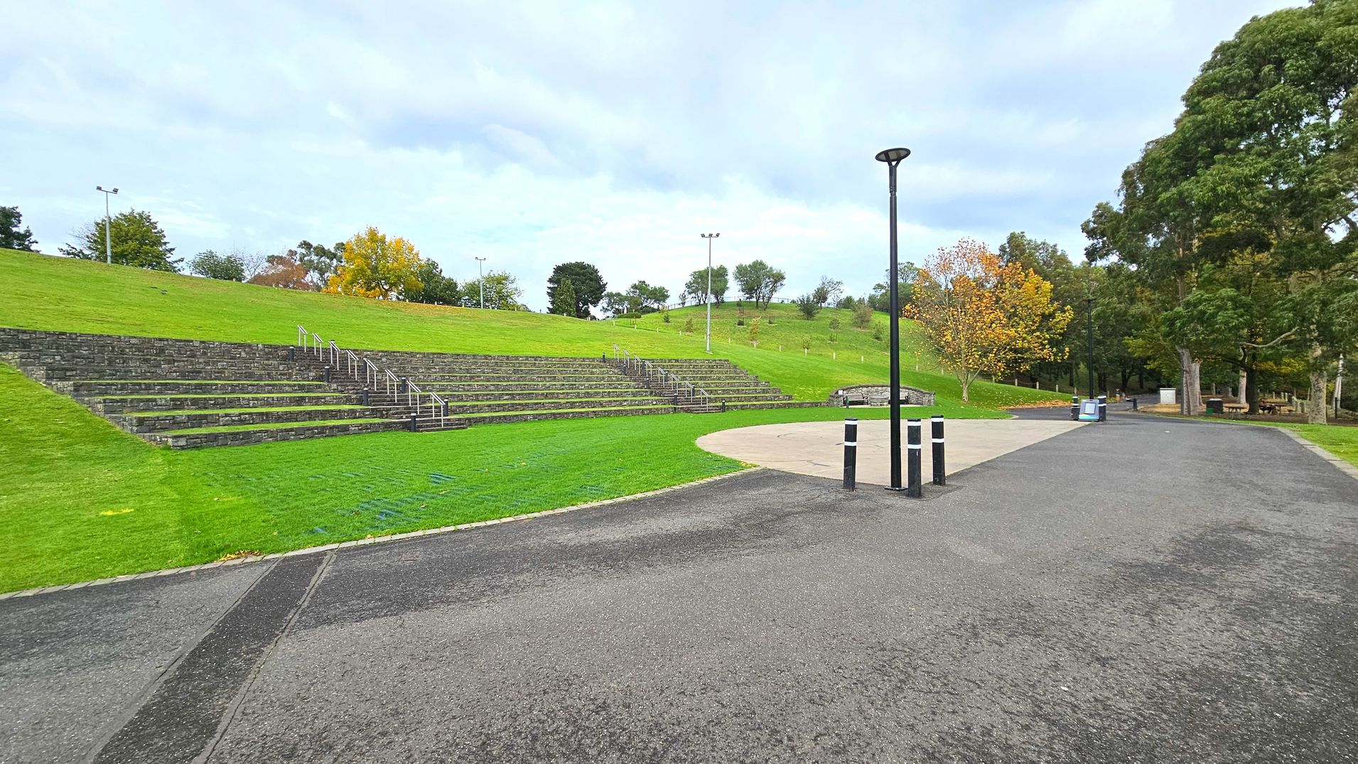 A park scene with a wide, paved path curving through the foreground, bordered by a grassy area on the left. Stone terraced seating is built into a grassy hill, with metal railings for access. A tall black lamp post stands near the path. Trees with autumn foliage are scattered around, and a few benches and bins are visible in the distance. The area feels open with a gentle slope upwards.