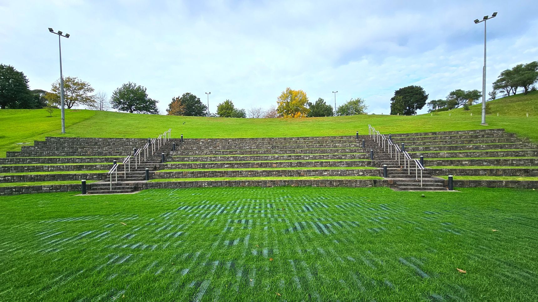 An outdoor amphitheatre with tiered stone seating arranged in a semi-circle on a grassy slope. The seating is divided by two sets of metal railings leading up the steps. The foreground features a well-maintained grass area with visible grid patterns. Tall floodlights are positioned on either side of the seating area. Trees are scattered along the top of the slope, providing a natural backdrop.