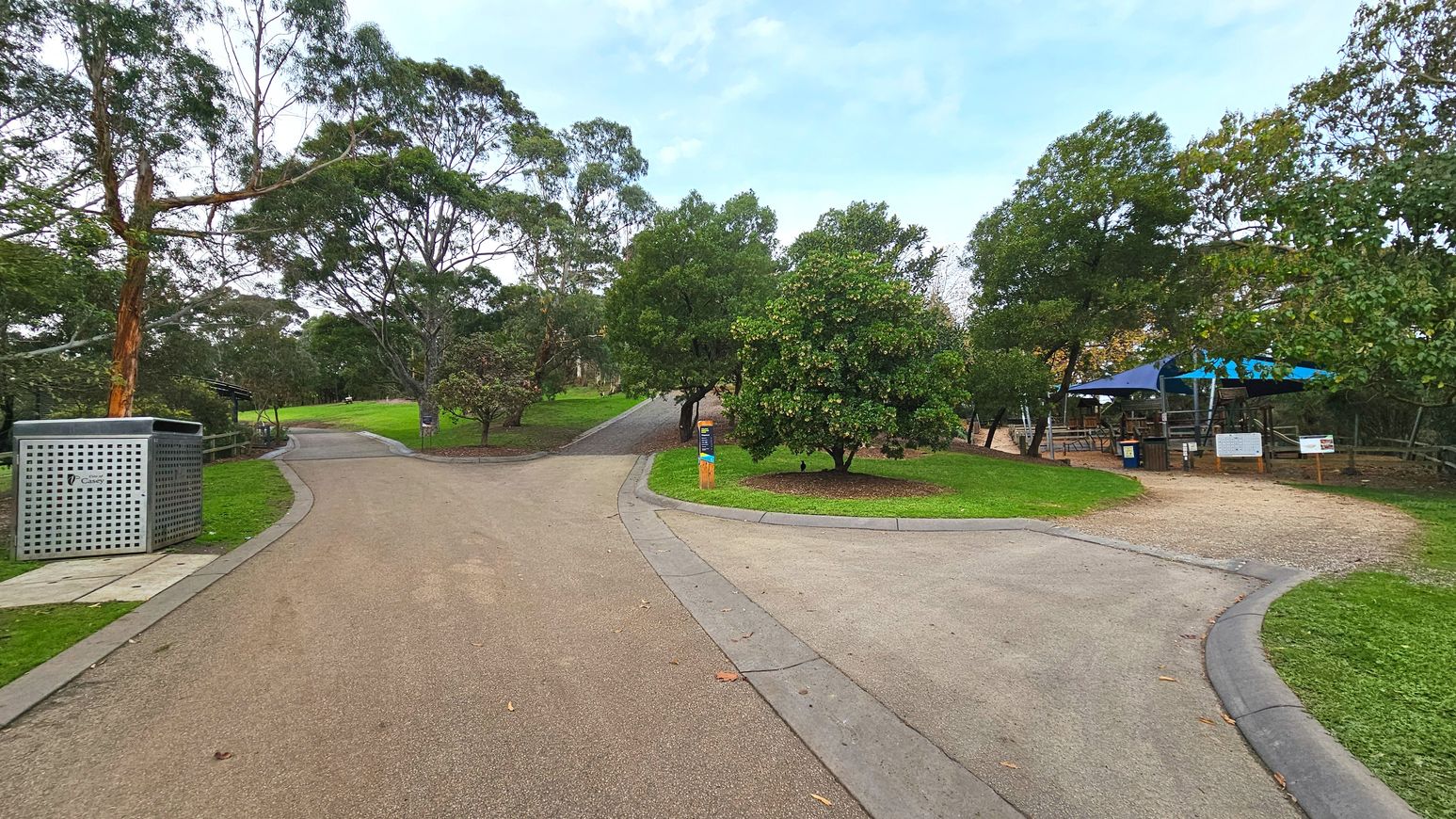 A park scene with a paved path splitting into two directions. On the left, the path curves gently uphill, bordered by a metal bin and a wooden fence. On the right, a gravel path leads to a seating area with tables under blue umbrellas. Trees and shrubs are scattered throughout, providing shade. A wooden post with a sign stands at the fork. The area feels open with greenery surrounding the paths.