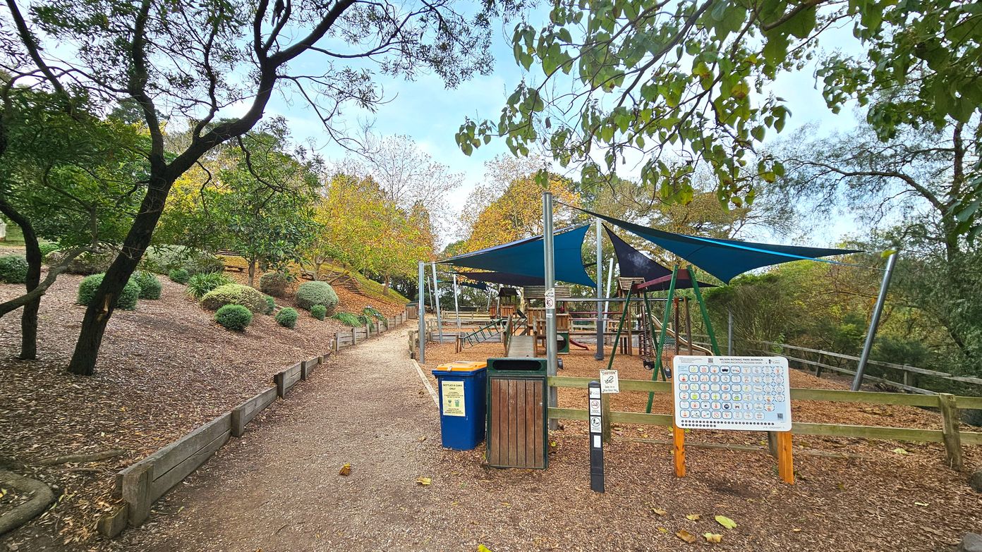 A playground area with a wooden climbing structure and swings is covered by blue triangular shade sails supported by metal poles. The ground is covered with wood chips. A path made of compacted dirt runs alongside, bordered by wooden planks. To the left, there are landscaped bushes and trees. A blue and a wooden bin are near the entrance, and a large sign with symbols is displayed on a wooden stand.
