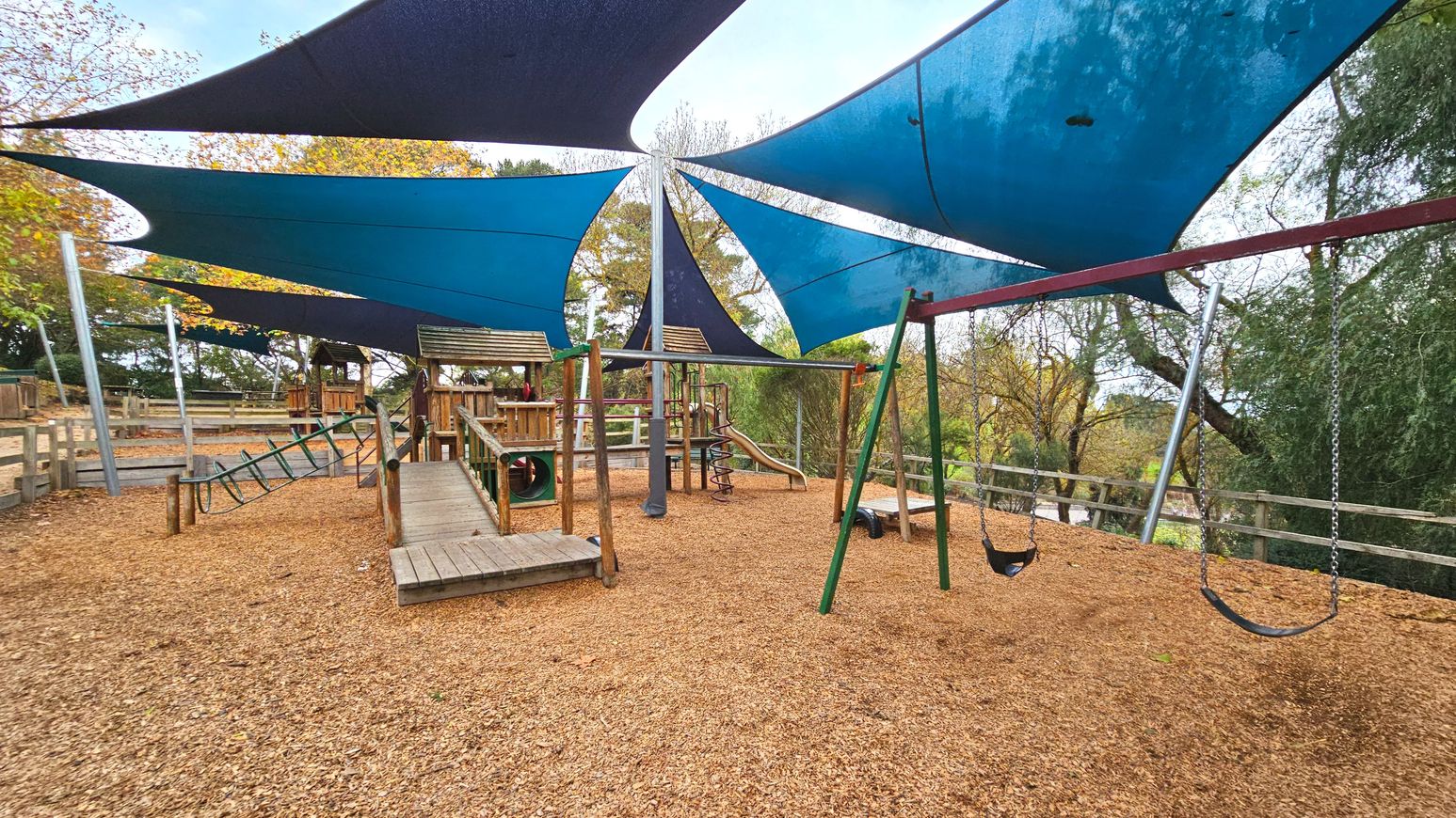 A playground with wooden structures including a slide, climbing frame, and swings, set on a surface covered with wood chips. Overhead, large blue and black triangular shade sails are stretched between metal poles. A wooden ramp leads up to a playhouse with a tunnel. The area is bordered by wooden fencing and surrounded by trees, creating a semi-enclosed space.