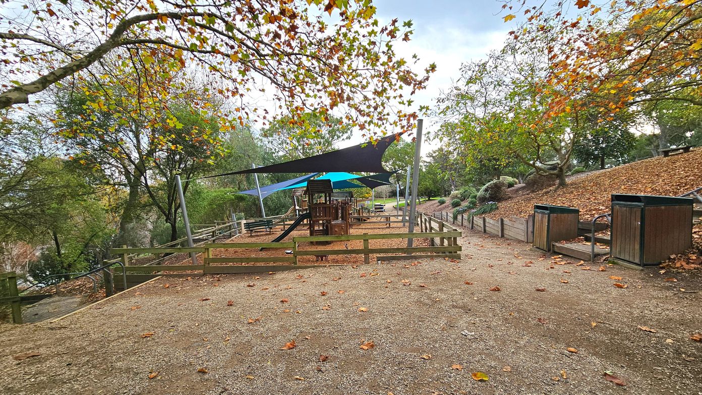 A playground area with a wooden climbing structure and slide is surrounded by a low wooden fence. Overhead, large triangular shade sails in black and blue provide cover. The ground is covered with wood chips, and the surrounding area has a gravel path with scattered autumn leaves. Wooden bins are placed along the path, and mature trees with colourful leaves frame the scene, creating a semi-enclosed space.
