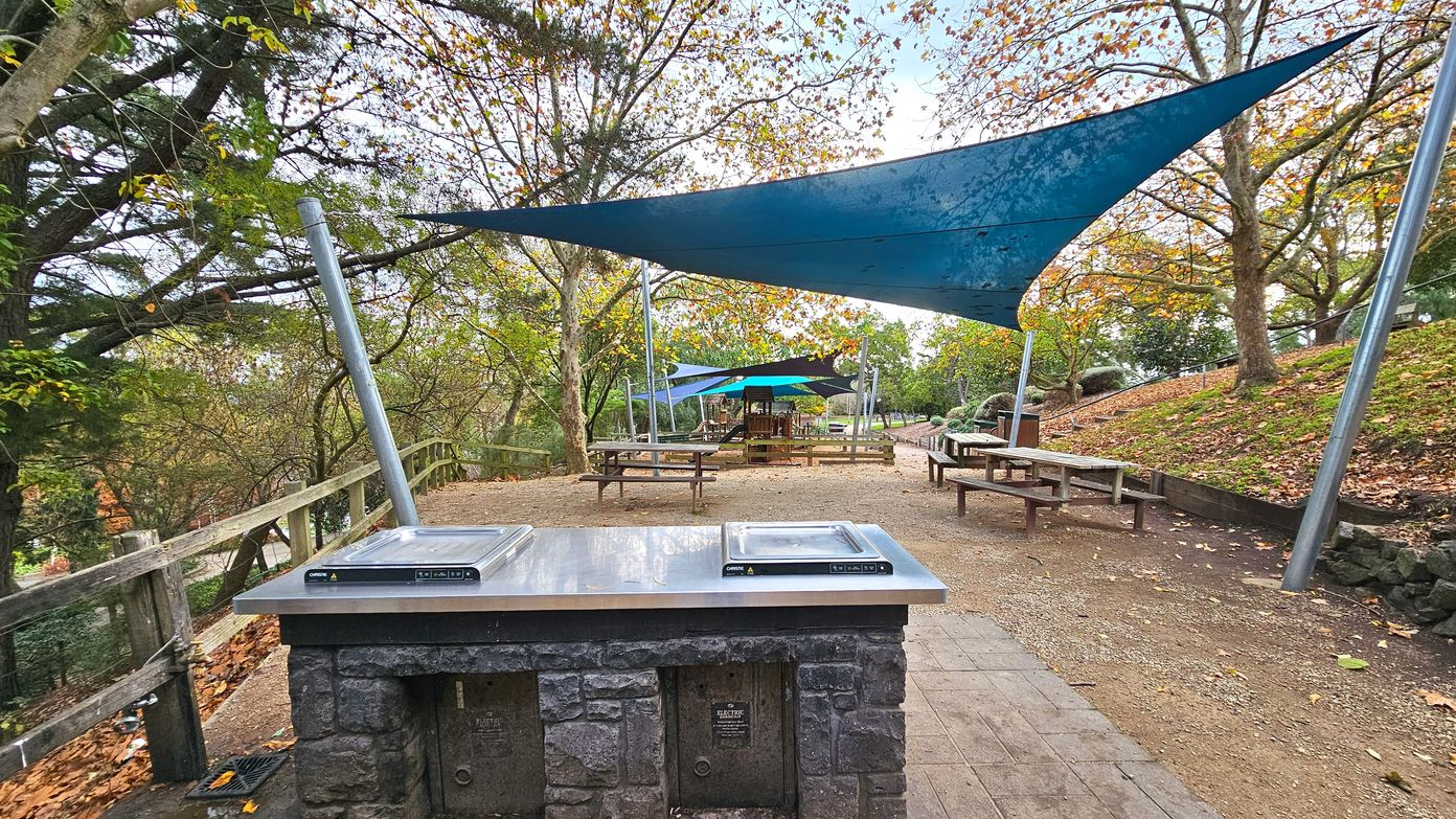 An outdoor picnic area features a stone barbecue with two stainless steel grills in the foreground. Several wooden picnic tables are scattered across the dirt and paved surface. Blue triangular shade sails are suspended above, supported by metal poles. A wooden fence runs along the left side, and trees with autumn leaves surround the area, creating a semi-enclosed space with a natural feel.