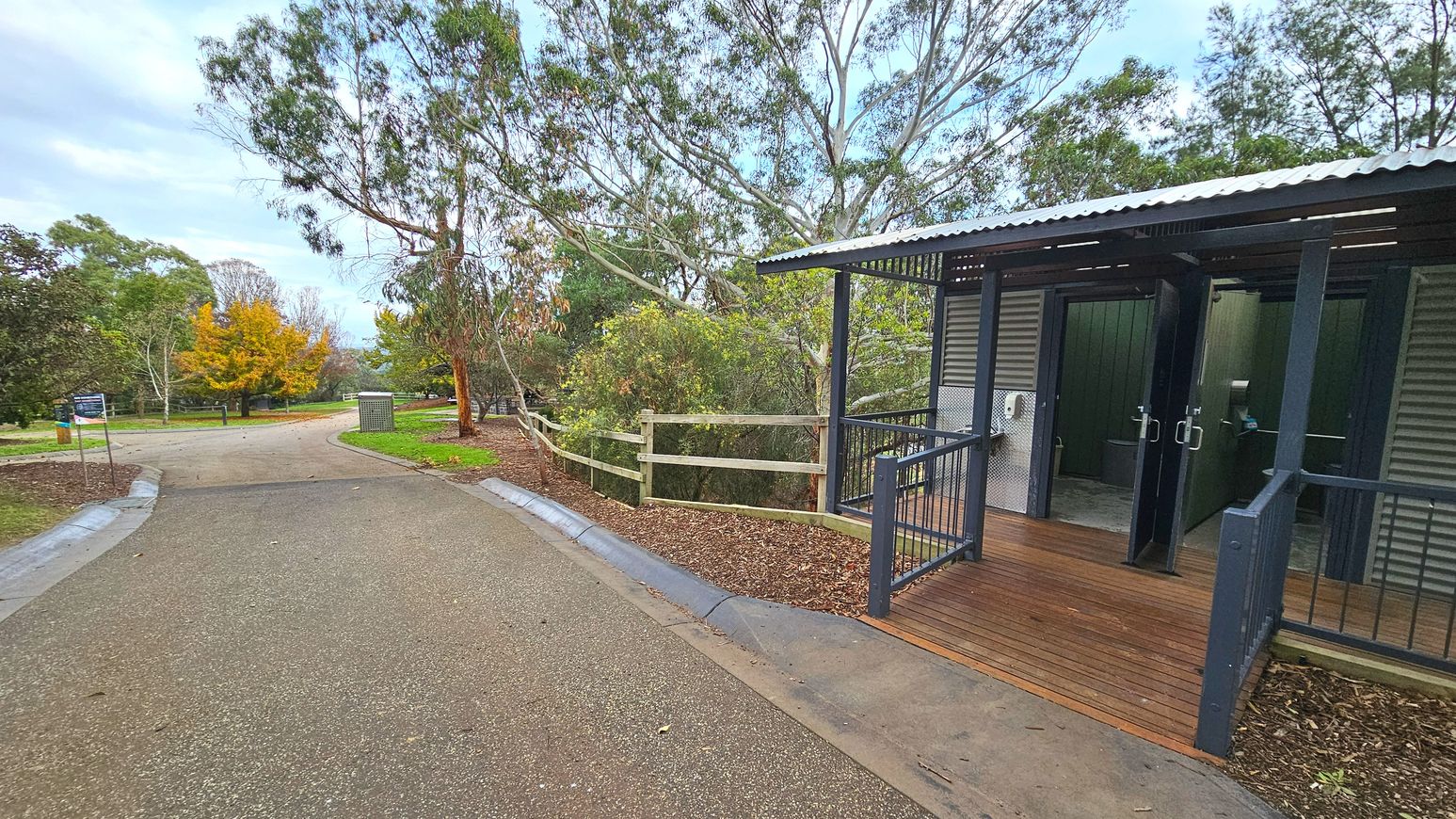 A public restroom facility with corrugated metal walls and a corrugated roof is situated on the right. It features a wooden ramp with metal railings leading to the entrance. The path in front is paved with a mix of asphalt and concrete, bordered by a low kerb. To the left, a dirt path curves into a park area with trees and a wooden fence. A signpost and a bin are visible along the path.