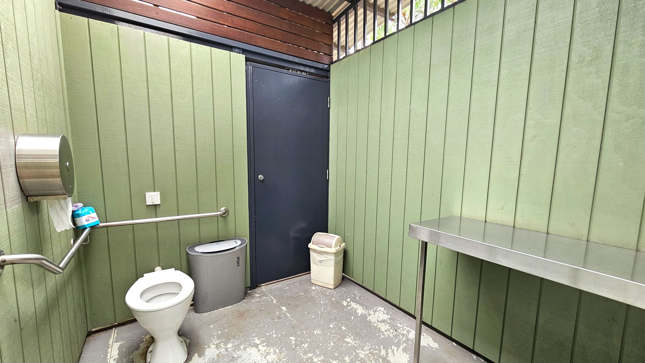 A public toilet with green panelled walls and a grey metal door. A white toilet with a metal grab rail is on the left, accompanied by a stainless steel toilet paper dispenser. A grey sanitary bin is beside the toilet. A beige waste bin is near the door. On the right, a stainless steel shelf is mounted on the wall. The floor is concrete with patches of wear, and the ceiling has wooden slats and metal bars.