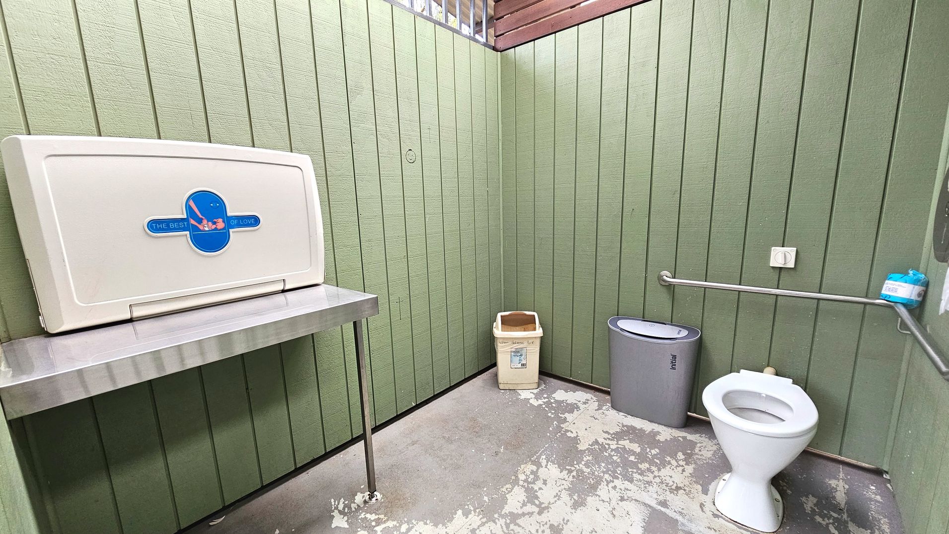 A public restroom with green vertical panelled walls and a concrete floor. On the left, a baby changing station is mounted above a stainless steel shelf. In the corner, a beige waste bin is placed. To the right, a white toilet with a metal grab bar beside it is visible. A grey sanitary bin is next to the toilet. A small shelf holds a container of wipes above the grab bar.