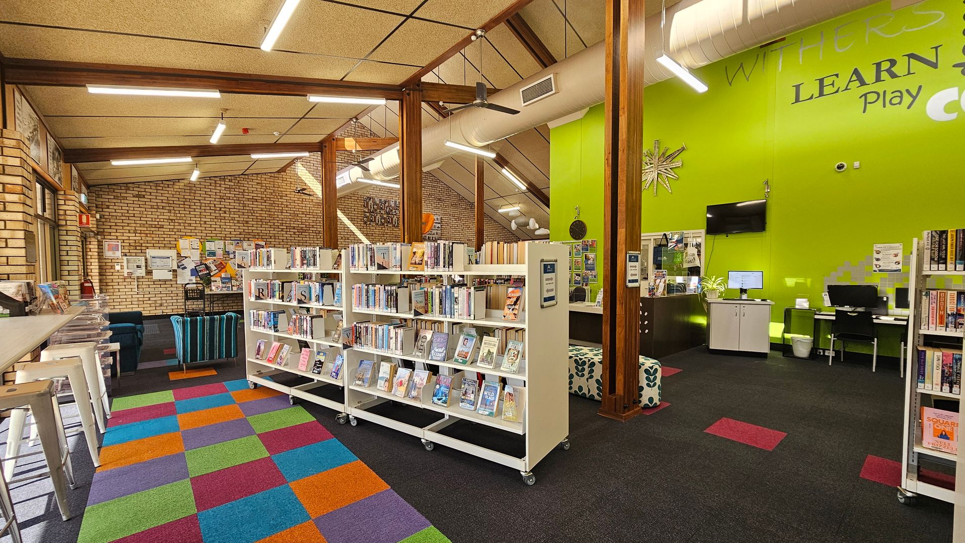 A library interior with a colourful, checkered carpet in the foreground. On the left, there are white bar stools and a brick wall with notices. In the centre, white bookshelves on wheels are filled with books. The right side features a bright green wall with a computer desk and a TV mounted above. The ceiling has exposed beams and fluorescent lights, with a visible air duct.