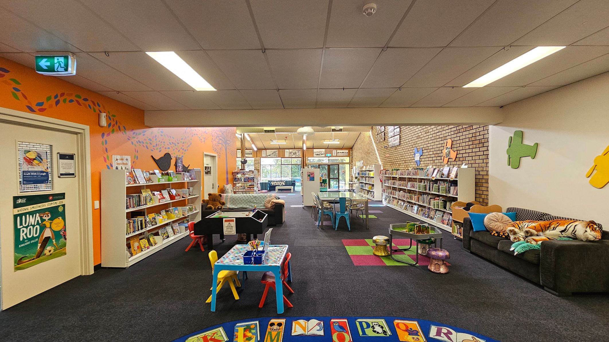 A colourful children's library area with an orange wall on the left featuring bookshelves filled with books and a poster. A small table with colourful chairs is in the centre, surrounded by a black carpet with an alphabet-themed rug. On the right, a dark sofa with plush toys, including a tiger, sits against a brick wall adorned with colourful plane cutouts. The ceiling has fluorescent lights, and the floor is carpeted with some green and red square patterns.