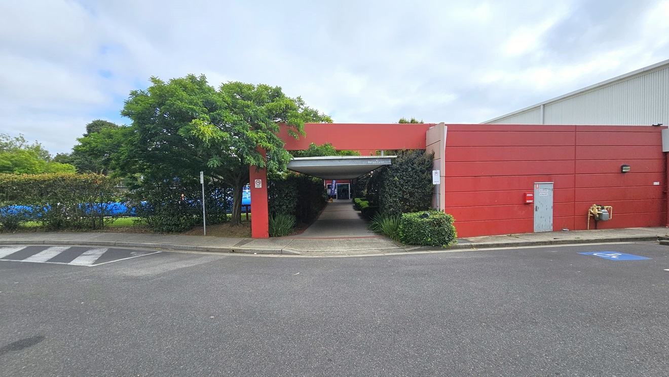 The facility comprises a large white corrugated iron warehouse design with an adjoining red concrete building. Next to the building is the car park, featuring accessible slots marked with a blue wheelchair sign. Yellow and black cars are parked, surrounded by tall trees. Additional parking is available on the left, and a step-free ramp leads to a lengthy open-air corridor.