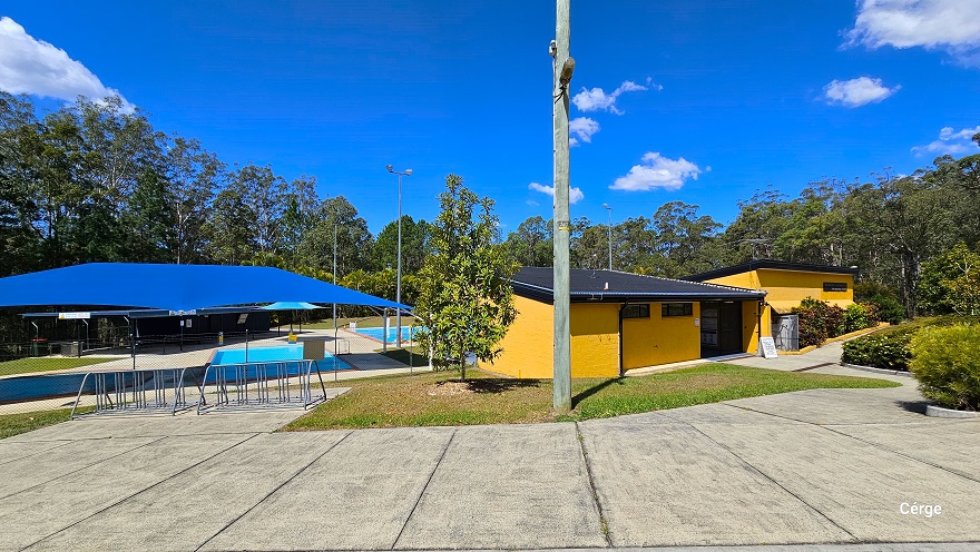 Concrete pathway near the two small toddler-wading outdoor pools and the administrative center with orange brick wall and dark grey corrugated roofing. On the pathway near the pool are two metal barriers. Beside it is a grass terrain with a medium-tall tree and a lamp post. A large blue sunshade covers the toddler pool. This image also shows the entrance to the orange brick admin building on the right side with two small windows on the upper left wall.