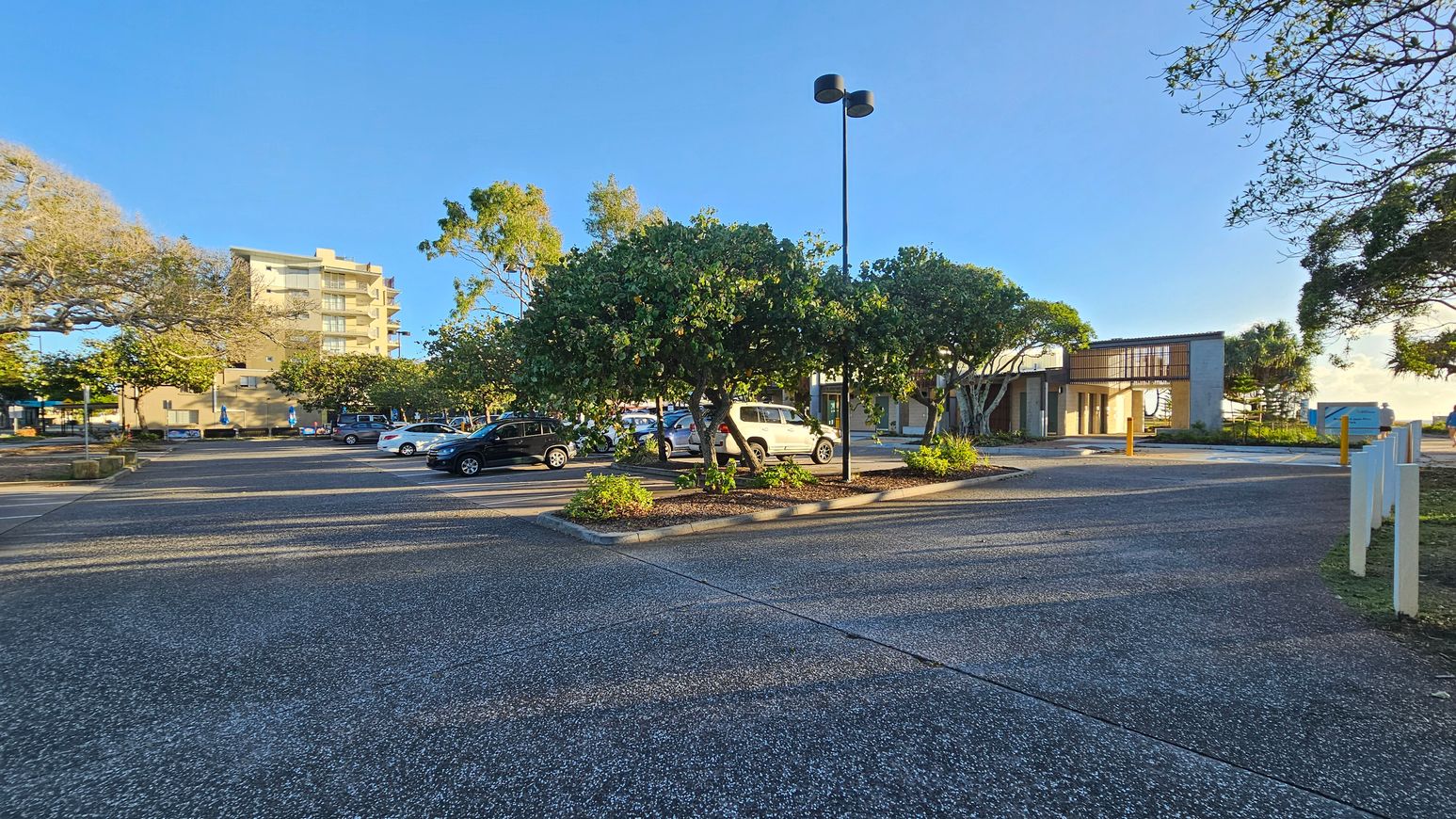 A car park with several vehicles is surrounded by trees and greenery. In the background, a multi-storey building is visible on the left, while a modern structure with a flat roof is on the right. The ground is paved, and there are patches of purple and pink hues on the surface. A tall streetlamp stands near the centre, and white posts line the right side of the image.