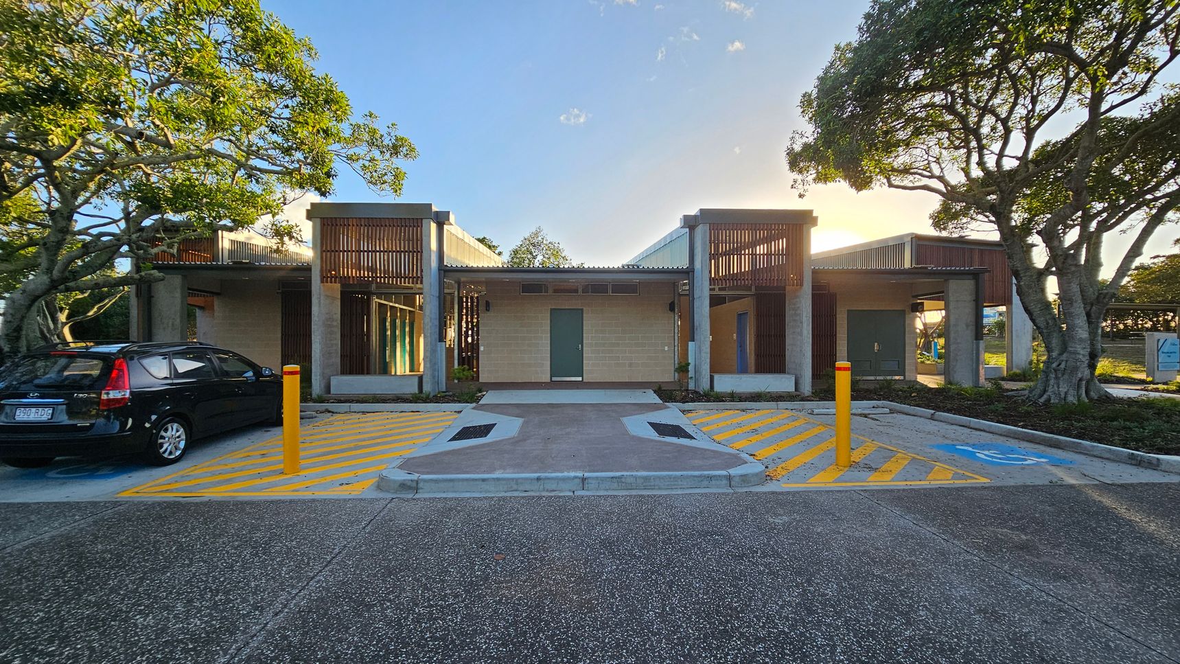 A modern building with a central entrance flanked by two sections featuring wooden slats. The foreground has a parking area with yellow-striped zones and a black car parked on the left. Two large trees frame the scene on either side, with sunlight filtering through the branches. The ground is a mix of textured concrete and asphalt, and there are yellow bollards near the entrance.