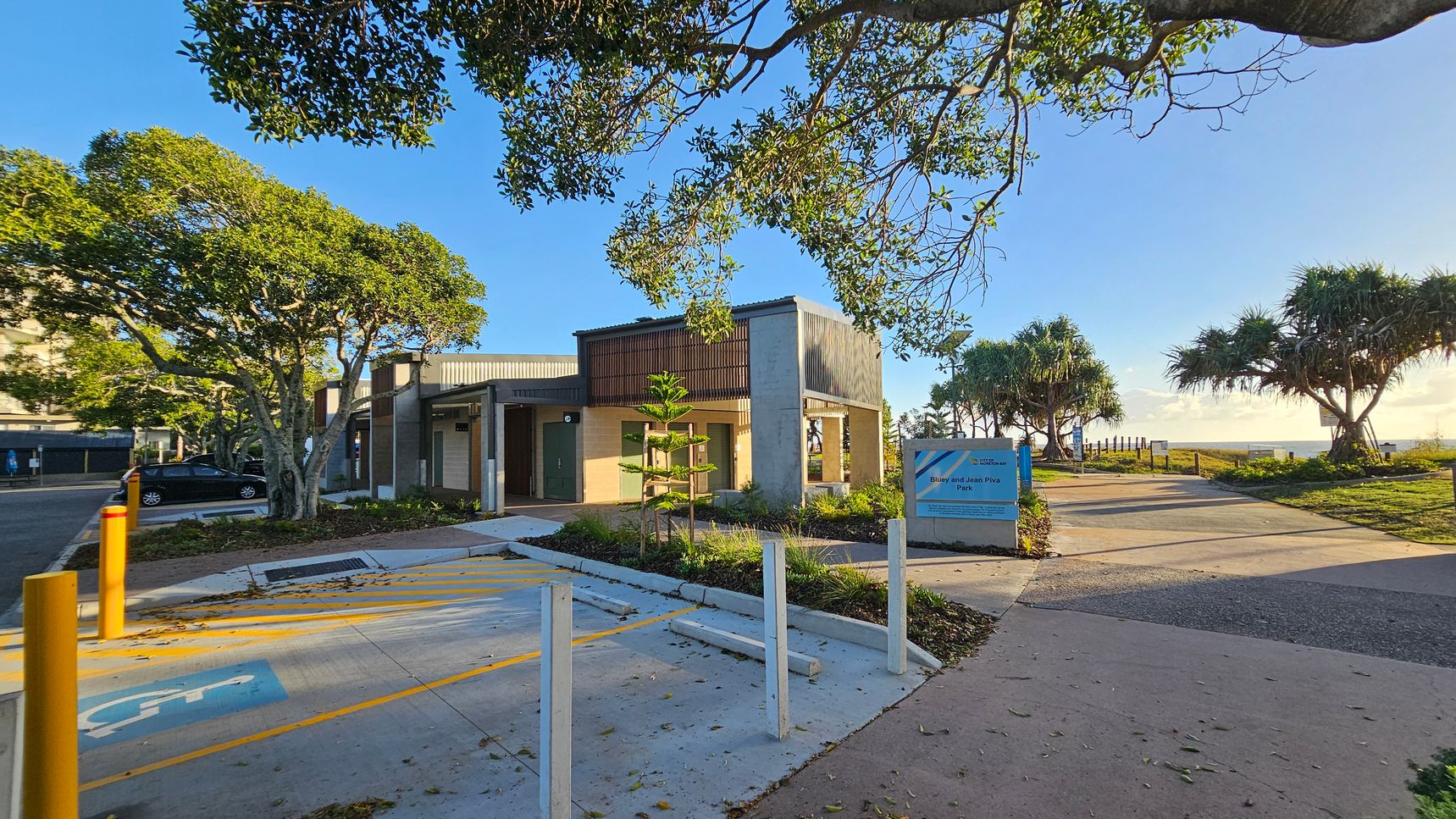 A small building with a modern design is surrounded by trees and greenery. On the left, a car is parked near a tree-lined street. In the centre, a pathway leads to the building entrance, with a sign indicating "Benny and Joan Piva Park" nearby. The right side features a paved path leading towards the ocean, bordered by more trees and grass. The sky is clear, and the area is well-lit by sunlight.