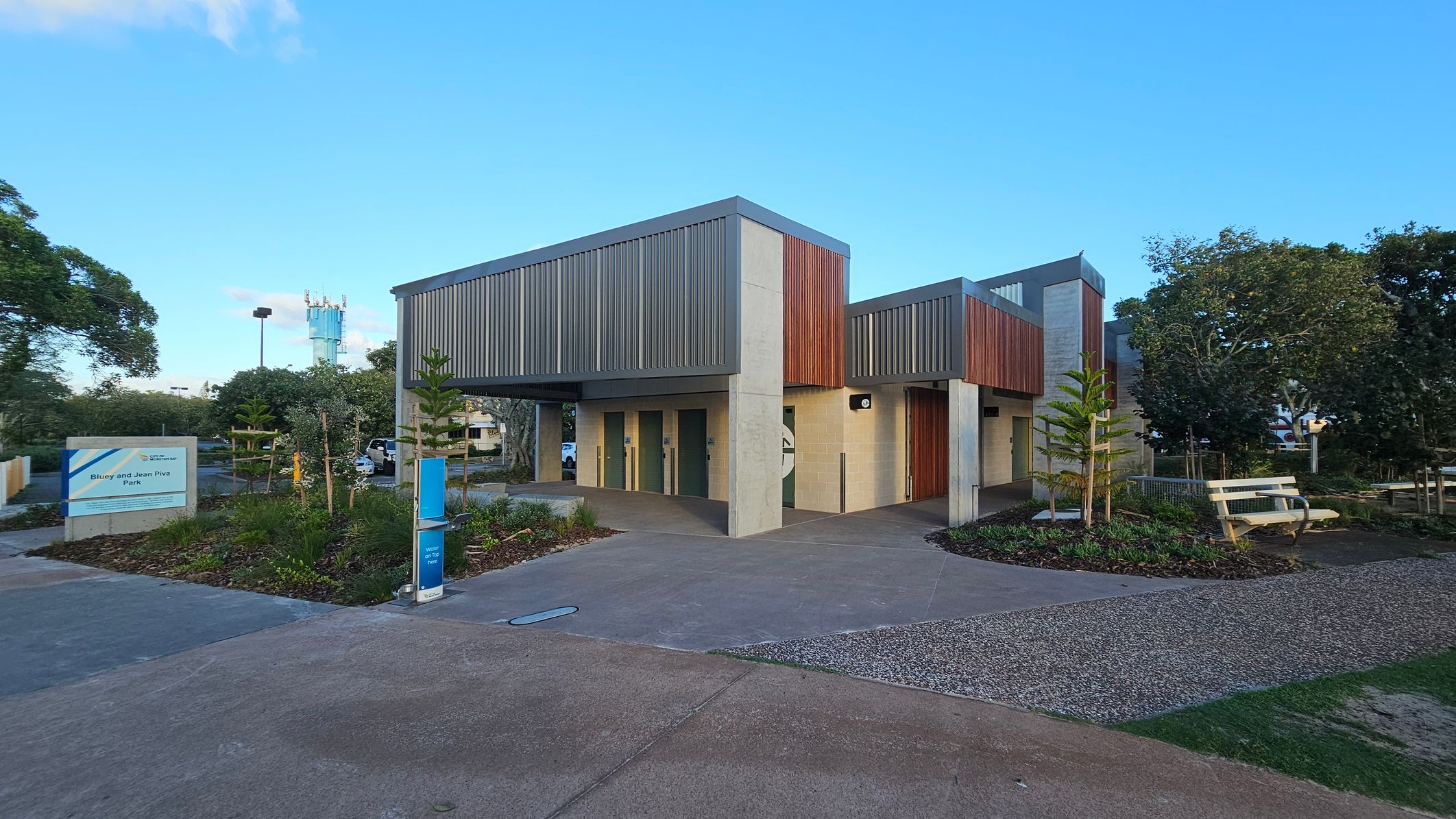 A modern building with a mix of vertical wooden and metal panels on the exterior. In the foreground, a paved path leads to the entrance, surrounded by landscaped gardens with small plants and trees. On the left, a sign reads "Biloy and Jean Riva Park." To the right, there are benches and more greenery. In the background, a tall structure is visible, partially obscured by trees. The sky is clear and blue.