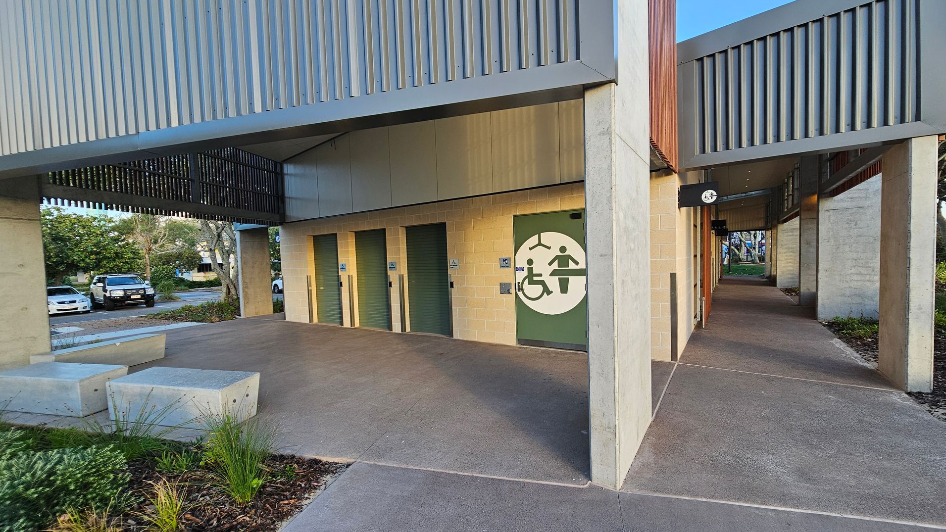A modern public restroom facility with a slatted metal roof and walls. On the left, there are three green doors, each with a sign indicating accessibility features. The rightmost door has a large symbol for disability access. A concrete path leads to the entrance, with a small garden and concrete benches on the left. In the background, there are parked cars and trees. The ceiling is open with metal slats, allowing natural light.