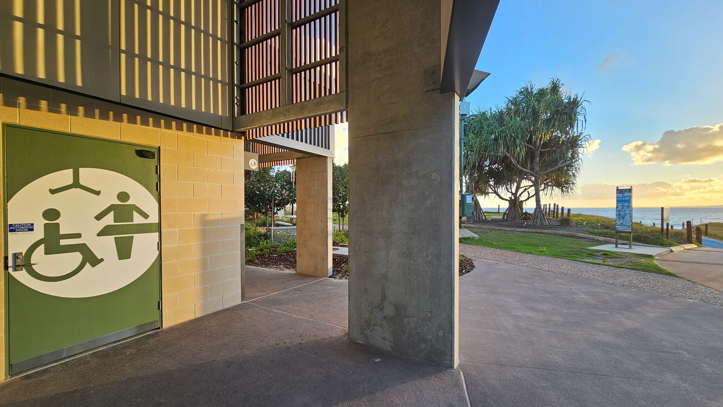 On the left, a building with a green door featuring a large accessibility symbol. The wall is made of light-coloured bricks. Above, there are wooden slats casting shadows. On the right, a concrete path leads towards a grassy area with trees and a view of the ocean. A signpost is visible near the water. The ground is a mix of concrete and grass, and the ceiling of the structure is not visible.