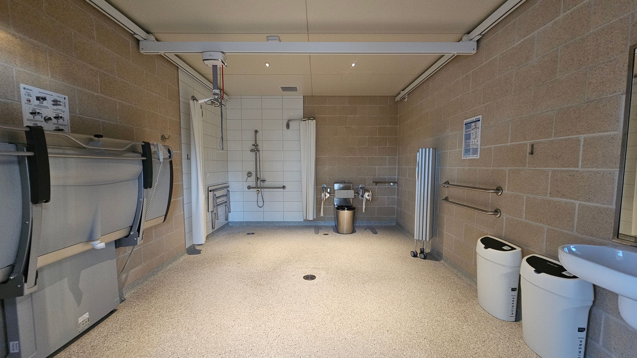 A spacious, accessible bathroom with beige tiled walls and a speckled floor. On the left, there's a foldable changing table and a ceiling track hoist. In the centre, a shower area with a curtain and grab bars. On the right, a toilet with support rails and a bin nearby. Two white bins are positioned against the right wall, and a sink is partially visible. The ceiling is plain with recessed lighting.