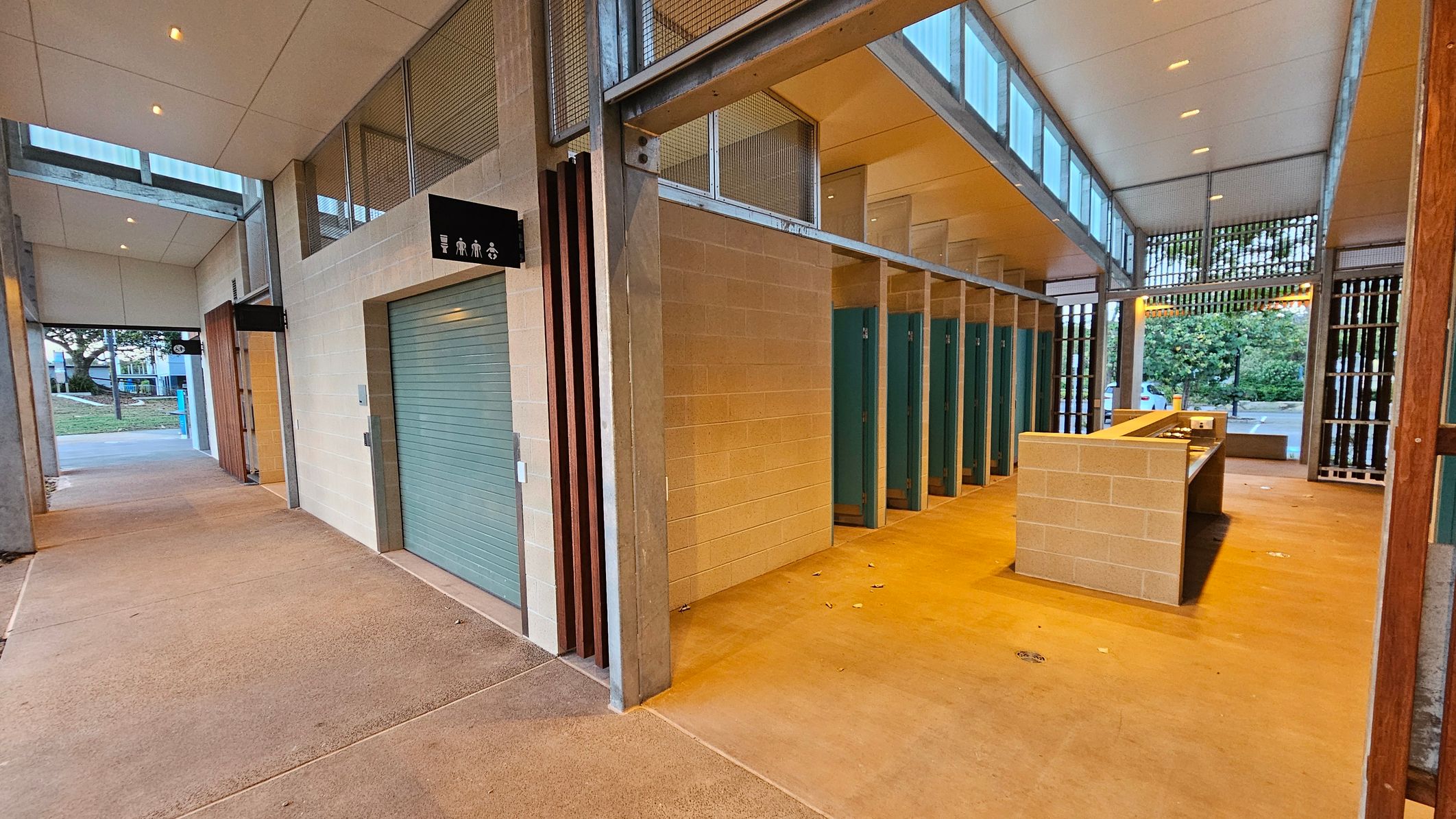 A public restroom with a beige tiled floor and a high ceiling with recessed lighting. On the right, there are several green stall doors and a central washbasin area with a countertop. The left side features a closed roller door and a sign with restroom symbols. The background shows an open area with trees and a car park visible through large windows and slatted wooden panels. The ceiling has a series of skylights allowing natural light in.
