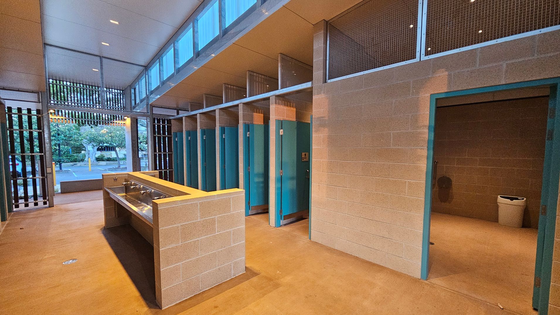 A public restroom with a row of blue-doored stalls on the right. In the centre, there is a long, rectangular sink with multiple faucets. The walls are made of light-coloured bricks, and the ceiling is high with small windows near the top. On the left, there is an open entrance with vertical slats, allowing a view of trees and a parking area outside. The floor is a smooth, light brown surface.