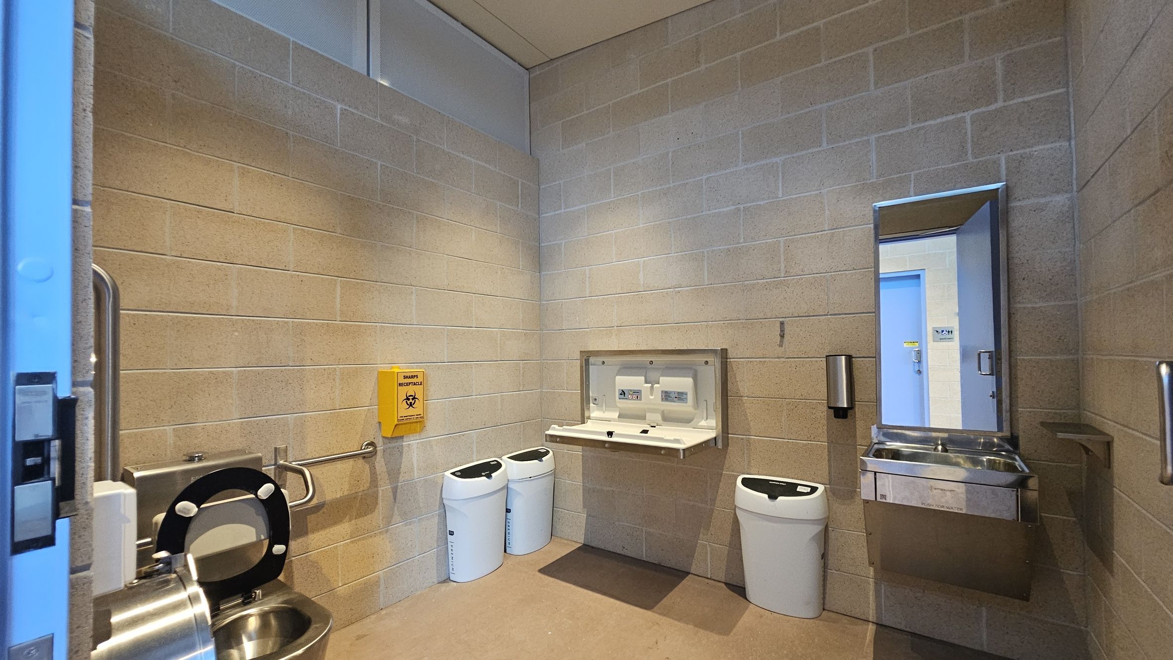 A public restroom with light-coloured brick walls. On the left, a stainless steel toilet with a black seat and a grab bar. A yellow sharps disposal box is mounted on the wall. In the centre, a baby changing station is folded down, with two white bins beneath it. On the right, a stainless steel sink with a mirror above it and a soap dispenser to the side. The floor is a light brown tile, and the ceiling is plain and light-coloured.