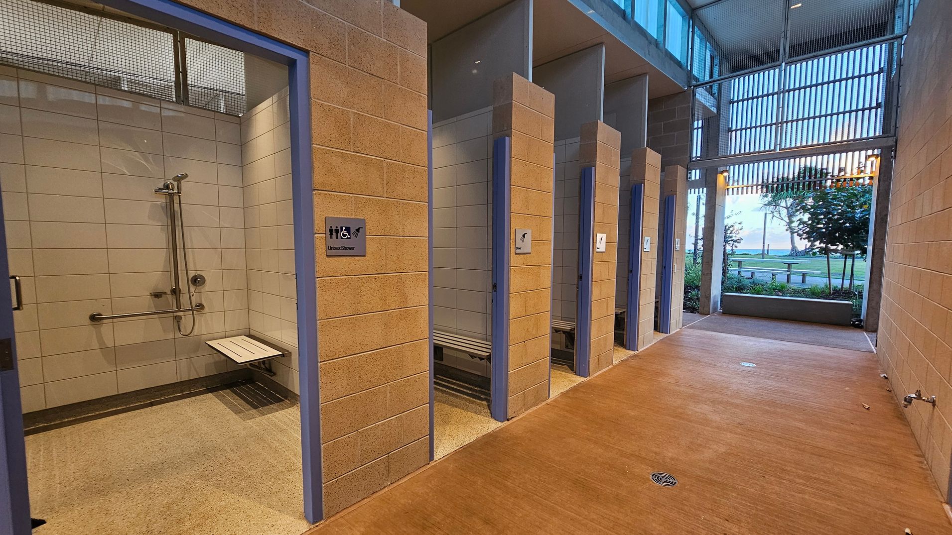 A row of shower stalls with beige brick walls and blue door frames. The first stall on the left is an accessible unisex shower with a foldable bench and grab bars. The floor is a light brown concrete with a drain. The ceiling is high with metal mesh panels. In the background, large windows and slatted panels allow a view of trees and a grassy area outside.