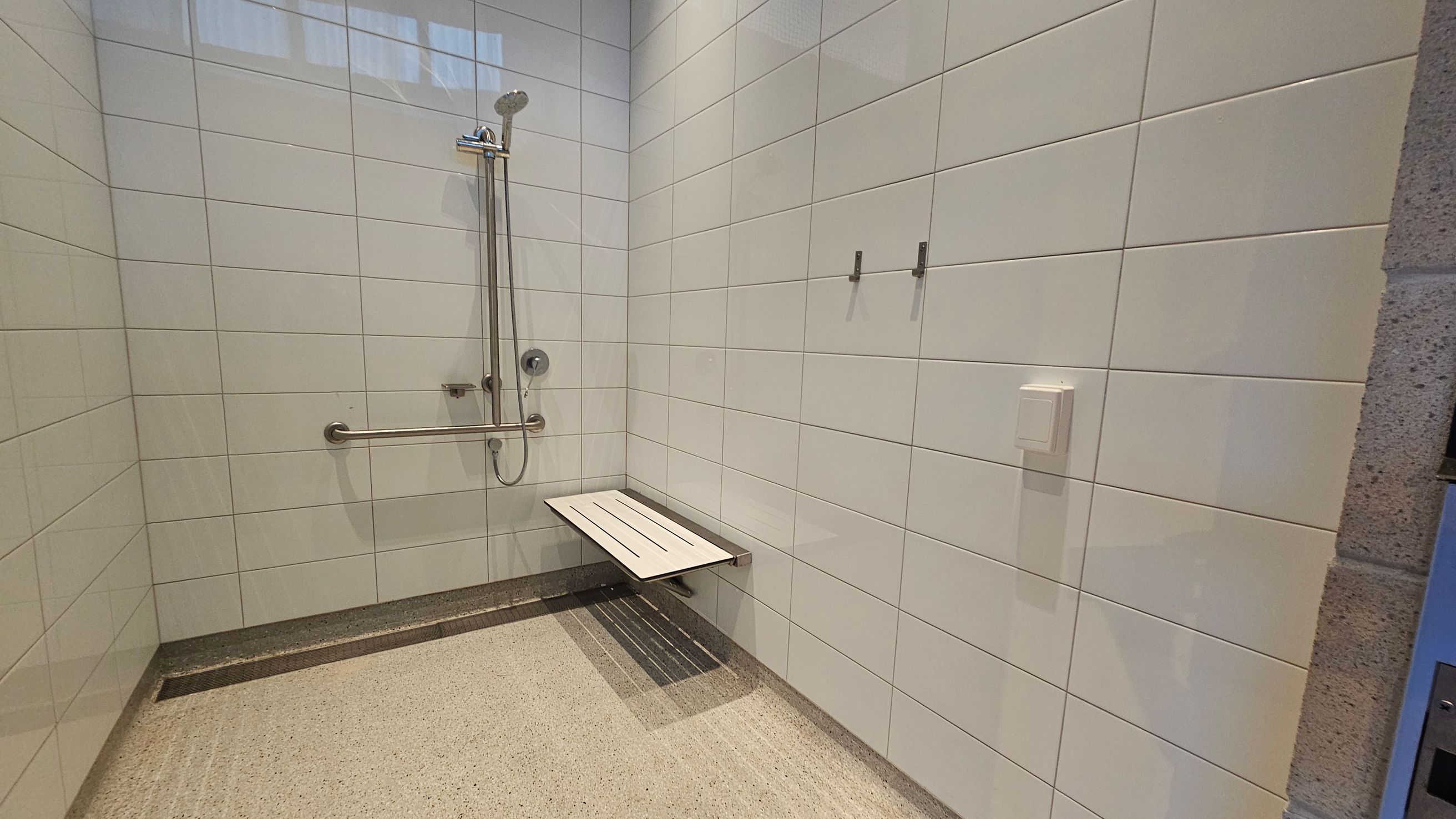 A shower room with light-coloured tiled walls. On the left, a showerhead is mounted on a vertical rail with an adjacent grab bar. Below, a foldable bench is attached to the wall. The floor is textured for safety. On the right wall, there are two hooks and a small rectangular fixture. The ceiling is not visible.