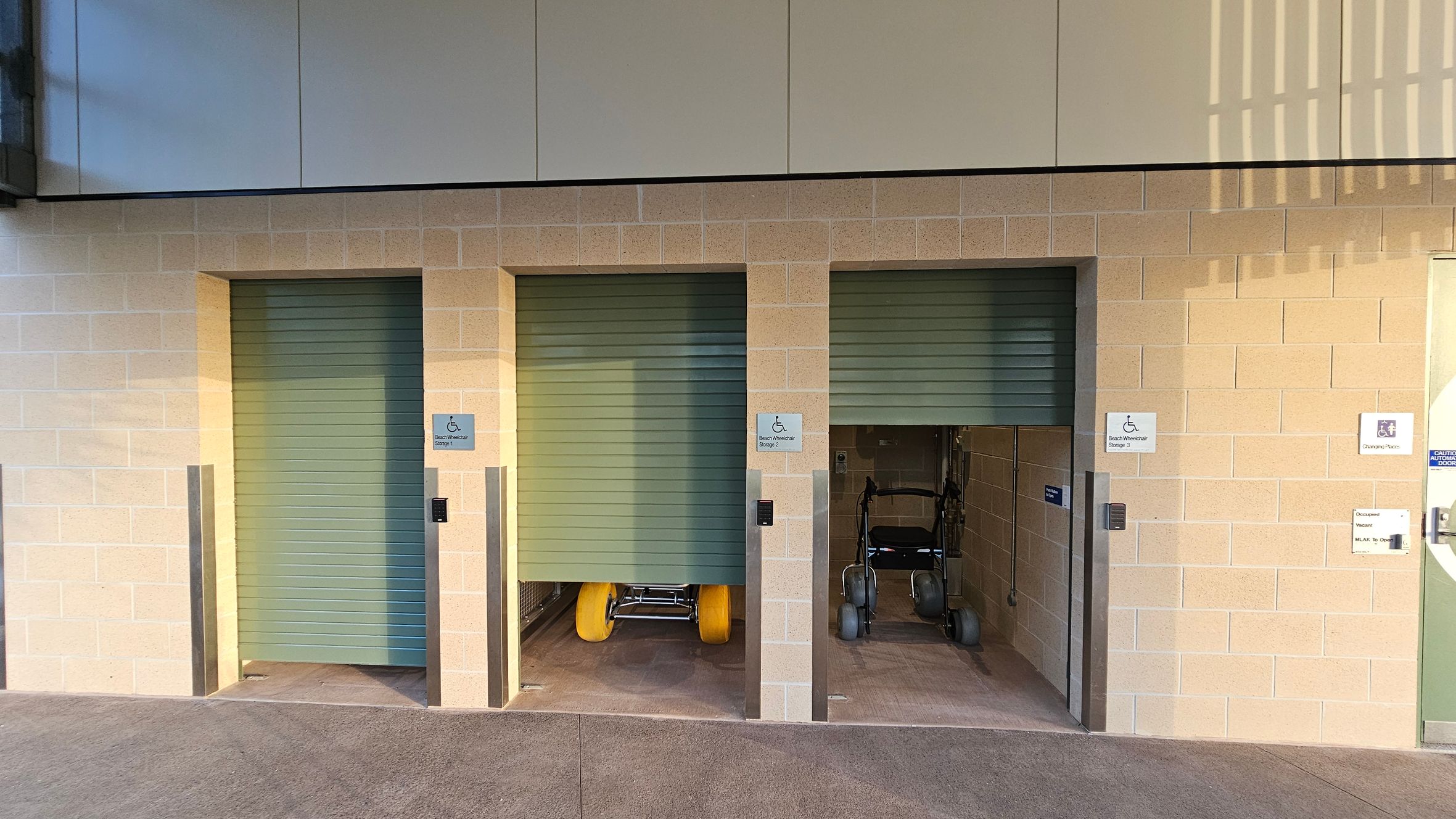 Three storage units with partially open green roller doors are set into a light brick wall. The left unit is closed, the middle unit reveals a beach wheelchair with large yellow wheels, and the right unit contains a standard wheelchair. Each unit has a sign indicating accessibility. The floor is concrete, and the ceiling is not visible. A sign on the right wall indicates a changing place.