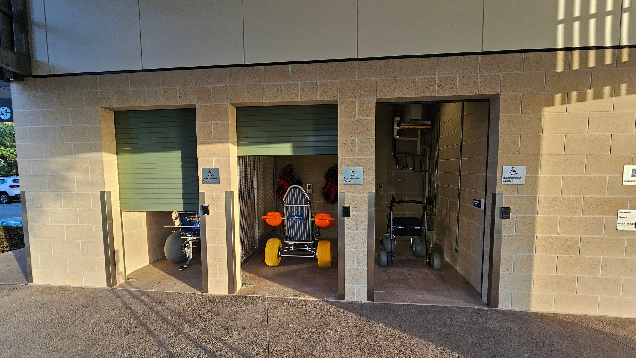 Three storage units with beige brick walls and green roll-up doors. The left unit contains a blue beach wheelchair with large white wheels. The middle unit has a beach wheelchair with yellow wheels and orange armrests. The right unit holds a standard wheelchair. Each unit has a sign indicating "Beach Wheelchair Storage." The floor is concrete, and the ceiling is not visible. Shadows from nearby structures are cast on the ground.