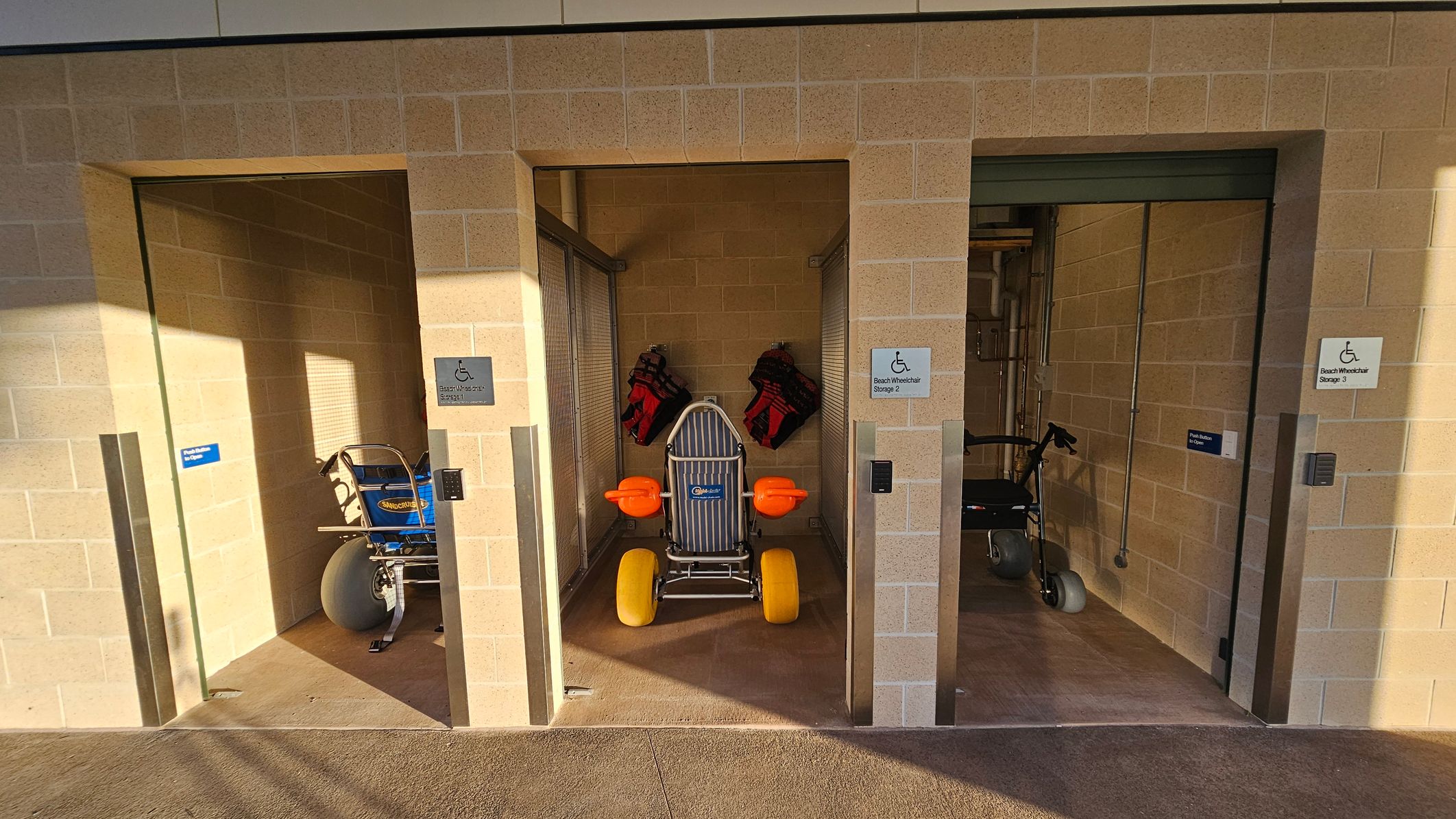 Three storage bays with beige brick walls. The left bay contains a blue beach wheelchair with large grey wheels. The middle bay has a beach wheelchair with yellow wheels and orange armrests, along with two red life vests hanging on the wall. The right bay holds a black wheelchair with grey wheels. Each bay has a sign indicating "Beach Wheelchair Storage" with a number. The floor is concrete, and the ceiling is not visible.