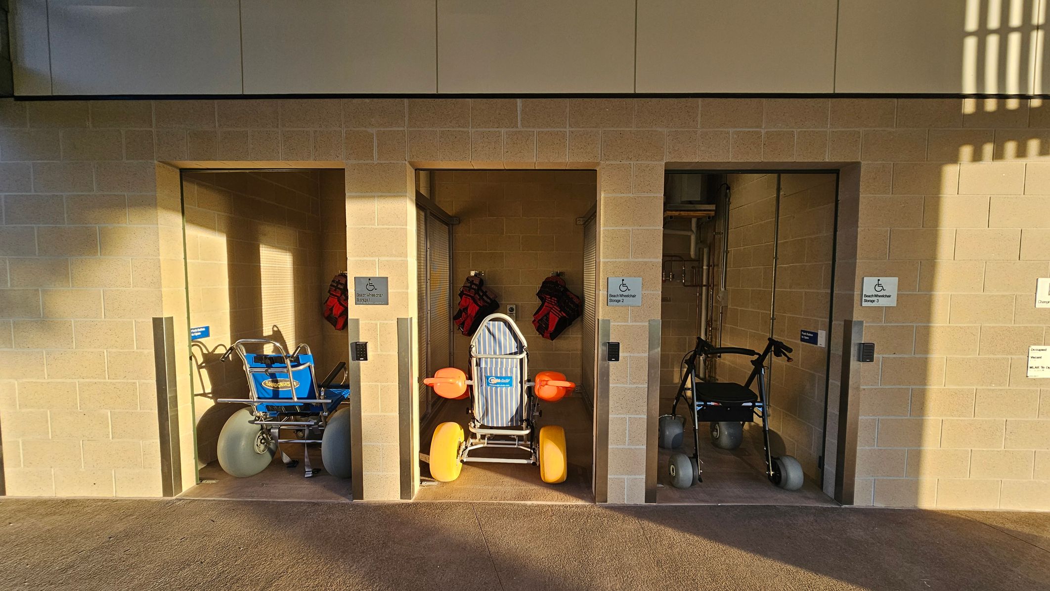 Three storage bays with beige brick walls. The left bay contains a blue beach wheelchair with large grey wheels. The middle bay has a striped beach wheelchair with bright orange armrests and yellow wheels. The right bay holds a walker with grey wheels. Each bay has a sign indicating "Beach Wheelchair Storage." The floor is a textured concrete surface, and the ceiling is not visible. Shadows from a structure above create patterns on the wall.