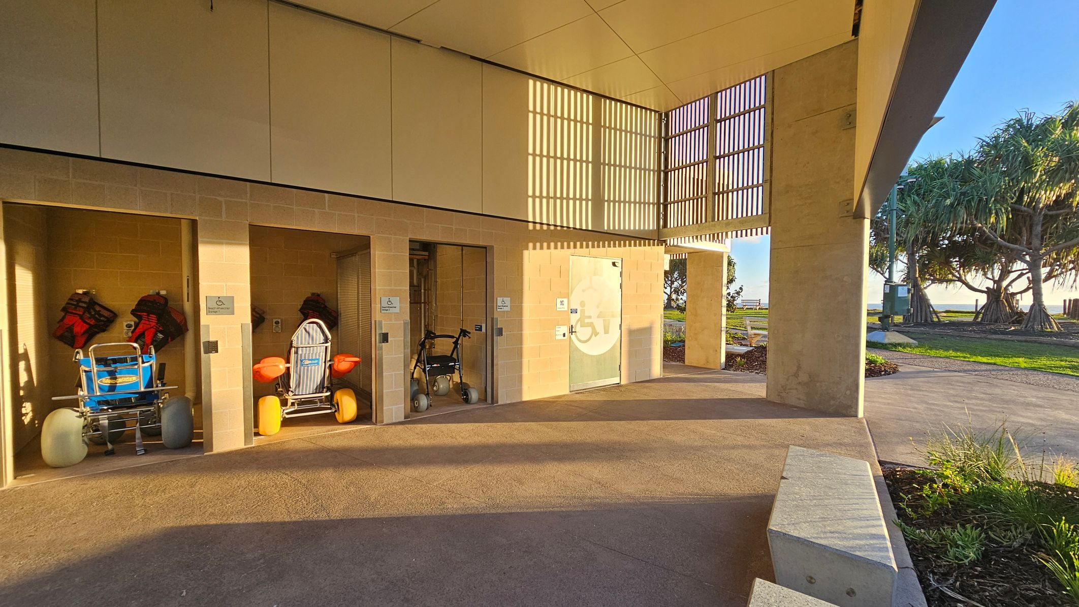 A row of storage units with open doors on the left, each containing beach wheelchairs with large wheels and life jackets. The units are made of light-coloured bricks. In the centre, a door with a wheelchair symbol. The floor is a mix of concrete and stone, and the ceiling is smooth with a grid pattern allowing light through. On the right, there are trees and a grassy area with a path leading towards the horizon.