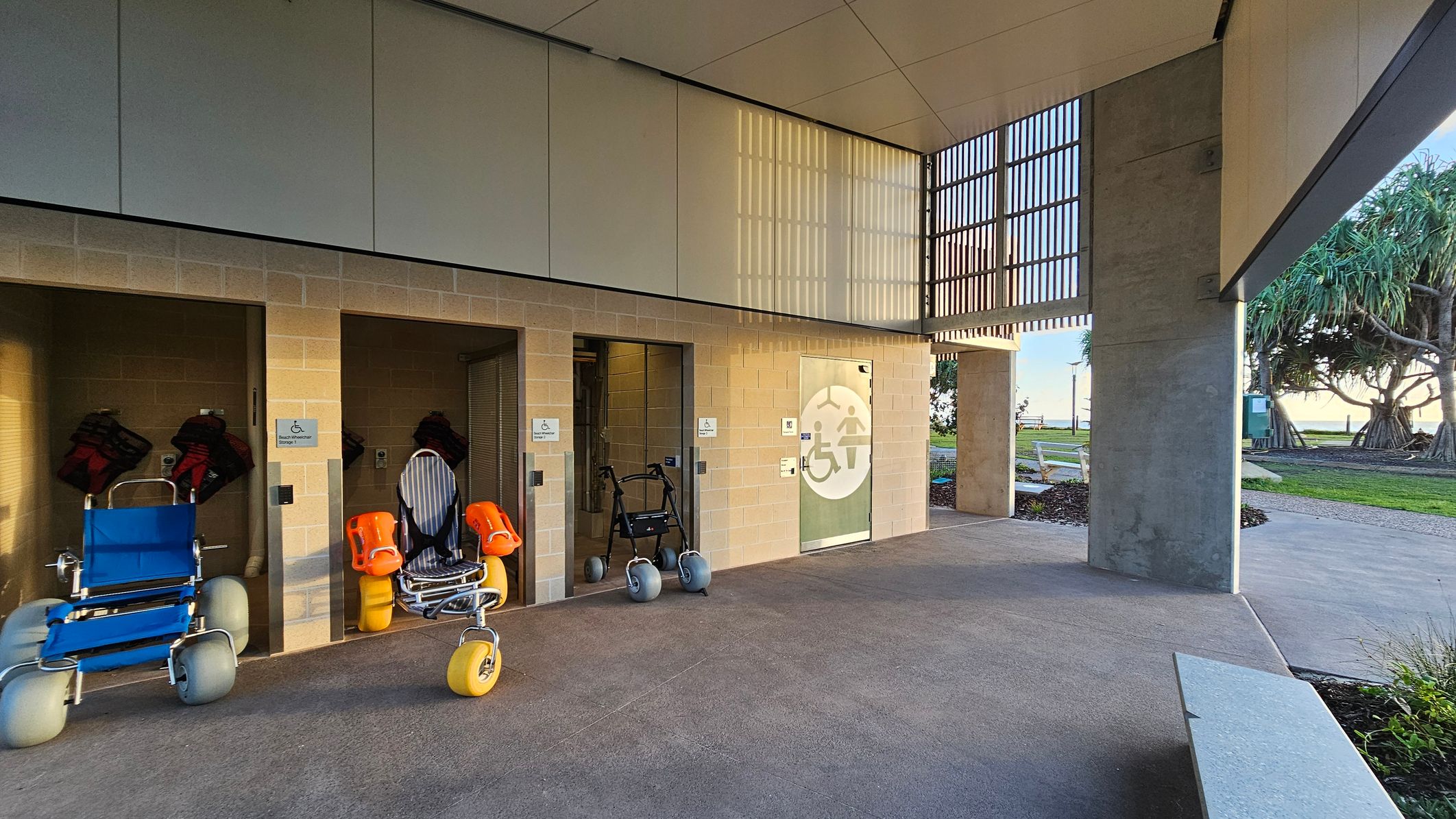 A sheltered area with three beach wheelchairs stored in individual alcoves on the left. Each alcove has a sign indicating accessibility. The floor is smooth concrete, and the ceiling is made of panels. On the right, there is a path leading to a grassy area with trees and a bench. The background shows a glimpse of the sea and sky through vertical slats.