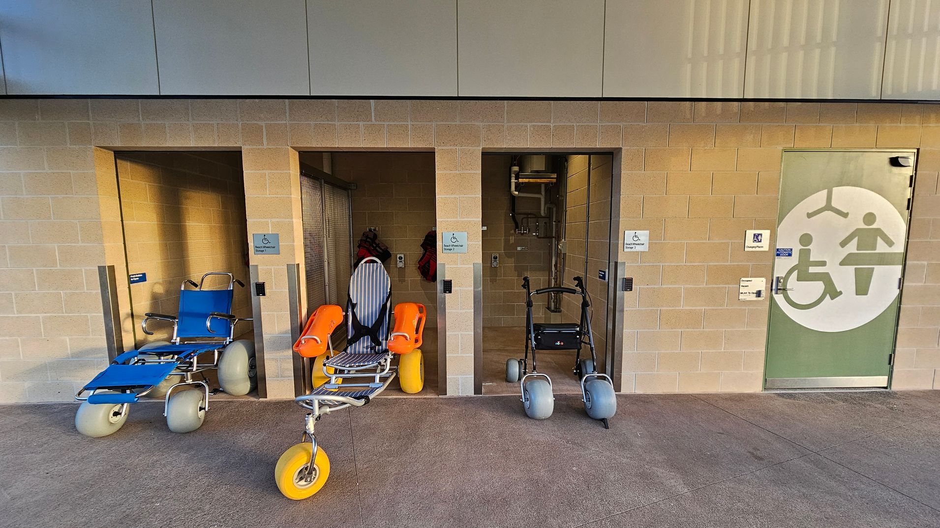 On the left, a blue beach wheelchair with large grey wheels is positioned in a recessed area. In the centre, an orange and grey beach wheelchair with yellow wheels is in another recessed space. To the right, a walker with large grey wheels is placed in front of a third recessed area. On the far right, a door with a large accessibility symbol is visible. The floor is concrete, and the wall is made of light-coloured bricks.