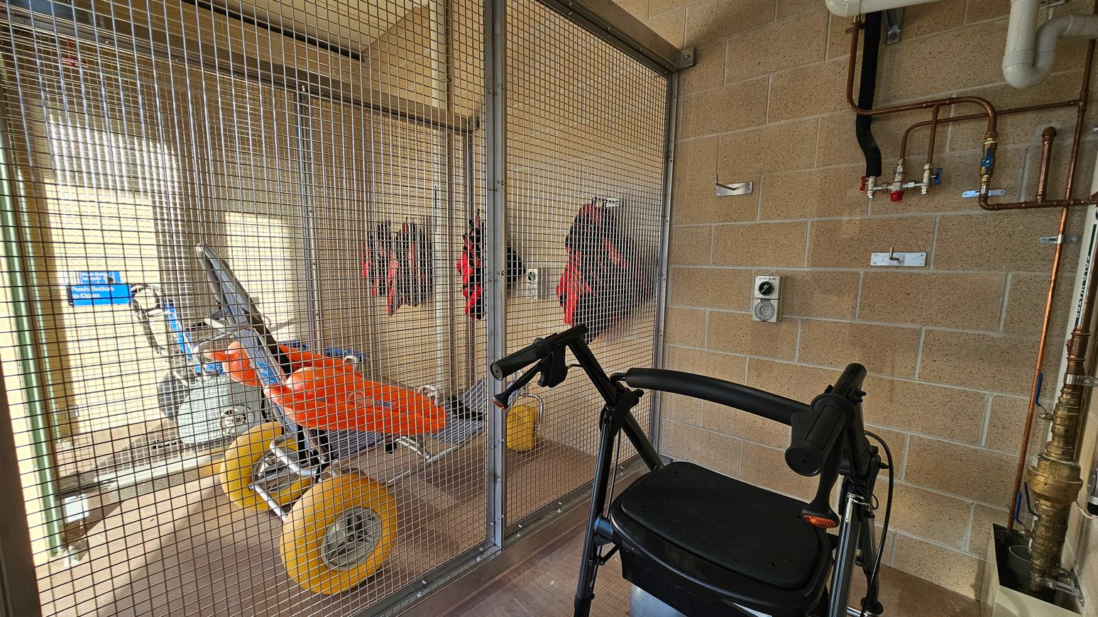 A room with a metal mesh enclosure on the left, containing an orange beach wheelchair with large yellow wheels. Several life jackets hang on the wall inside the enclosure. In the foreground, a black walker is positioned near the mesh. The right side features a brick wall with exposed pipes and valves. The floor is tiled, and the ceiling is not visible.