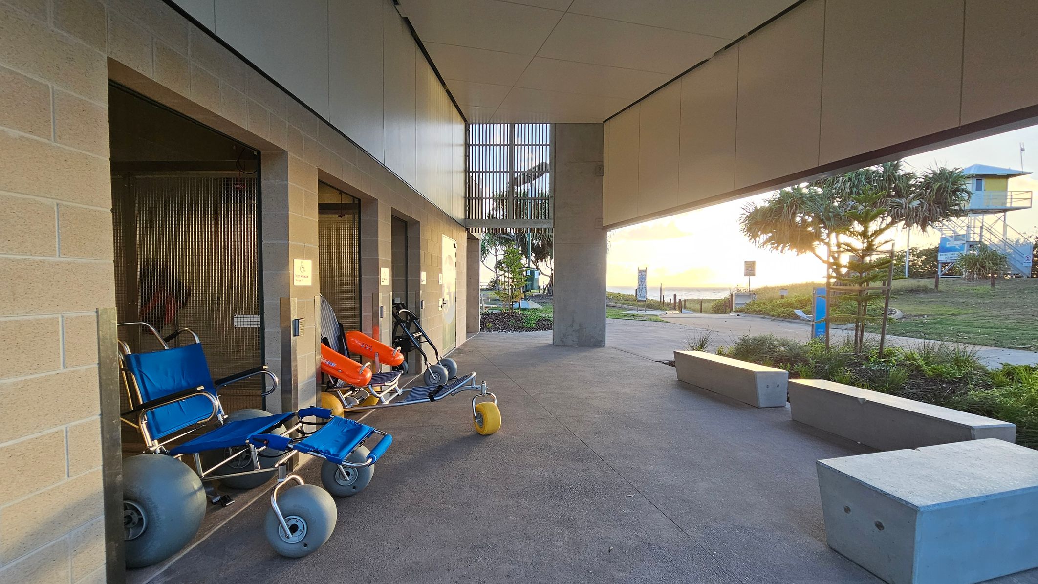 On the left, there are two beach wheelchairs with large grey wheels, one blue and one orange, positioned against a brick wall. In the middle, a concrete path leads towards the background, where a grassy area and a small tree are visible. On the right, there are two concrete benches and a view of the ocean in the distance. The ceiling is flat and modern, with a section of vertical slats allowing light through. The sun is setting, casting a warm glow over the scene.