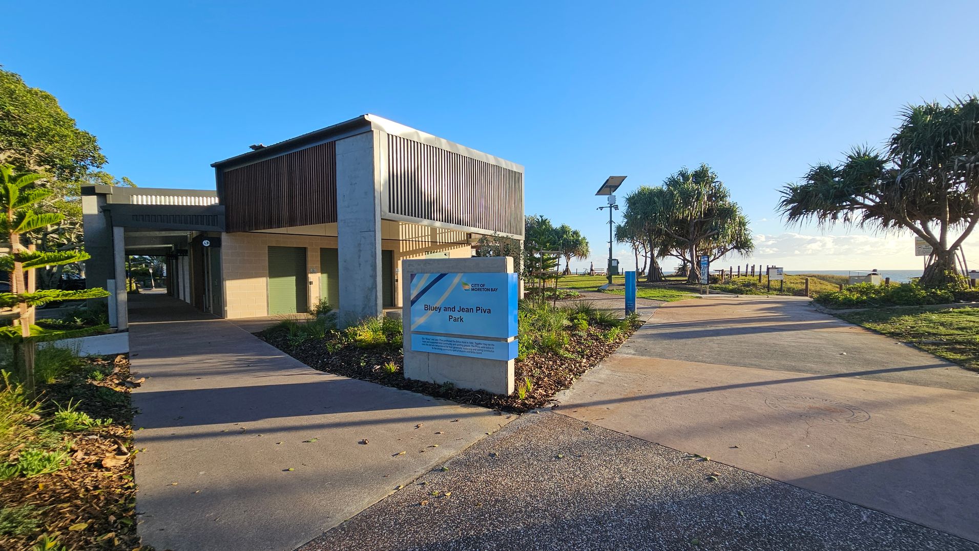 A modern building with vertical slats on the left, featuring a covered walkway. In front, a sign reads "Bluey and Jean Piva Park." A paved path leads to the right, bordered by grass and trees, including a few palm-like trees. A solar-powered light pole is visible along the path. The background shows a grassy area leading to a body of water, with a clear blue sky above.