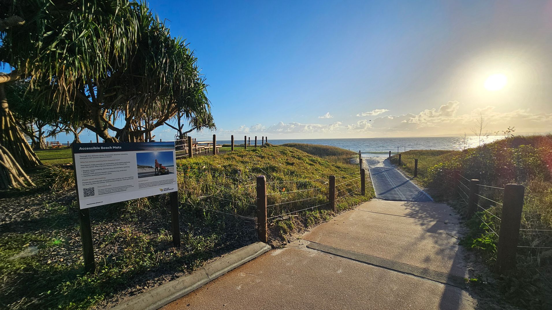 A pathway leads towards the ocean, with the sun low on the horizon. On the left, a sign about accessible beach mats is positioned near a cluster of large, leafy trees. The path is bordered by grass and low wooden posts with wire fencing. The ocean is visible in the background, reflecting the sunlight. The sky is clear with a few clouds near the horizon.