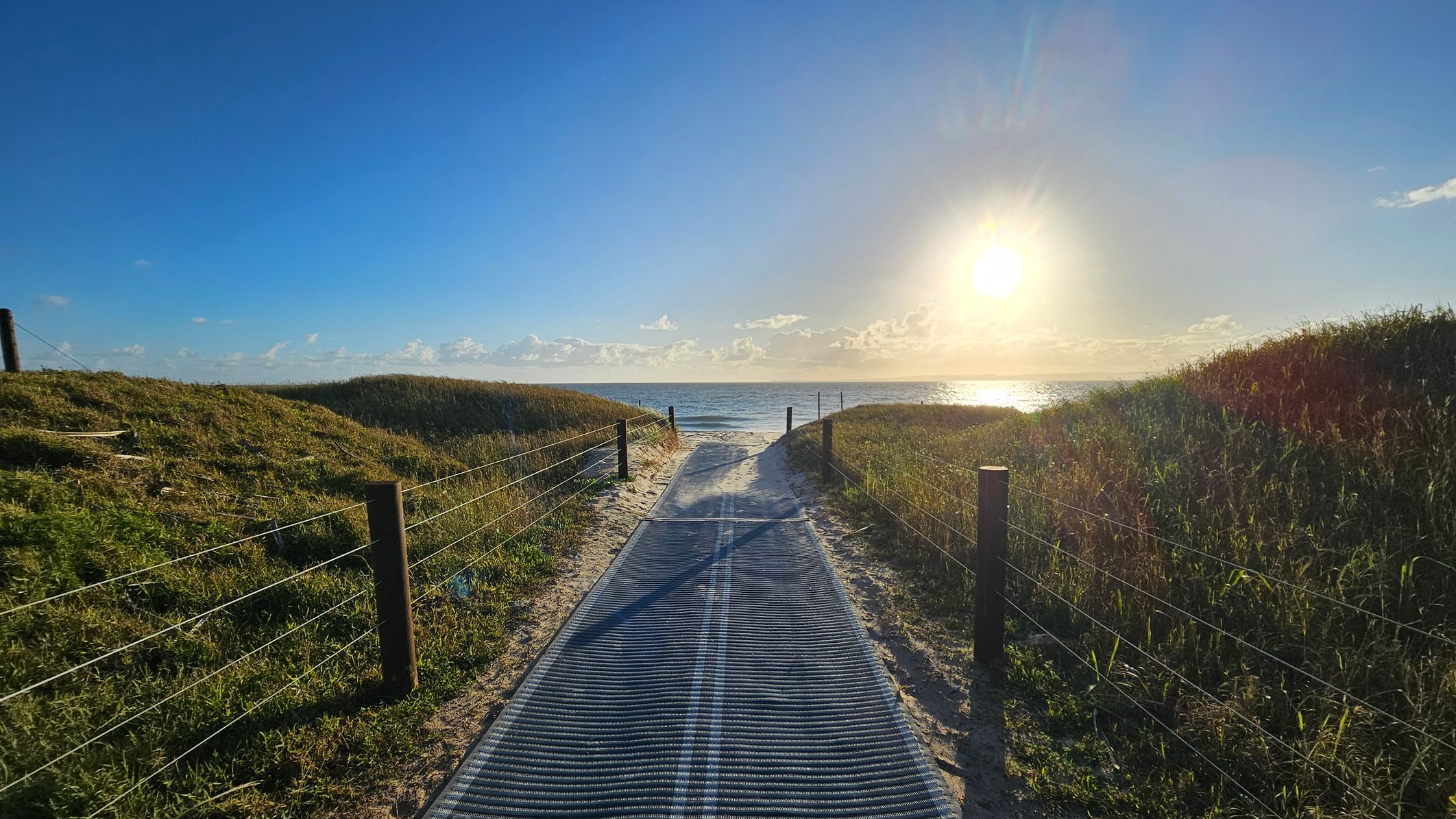 A sandy path leads towards the sea, flanked by grassy dunes on both sides. Wooden posts with wire fencing line the path. The sun is low in the sky, casting a bright reflection on the water. The sky is clear with a few clouds near the horizon.