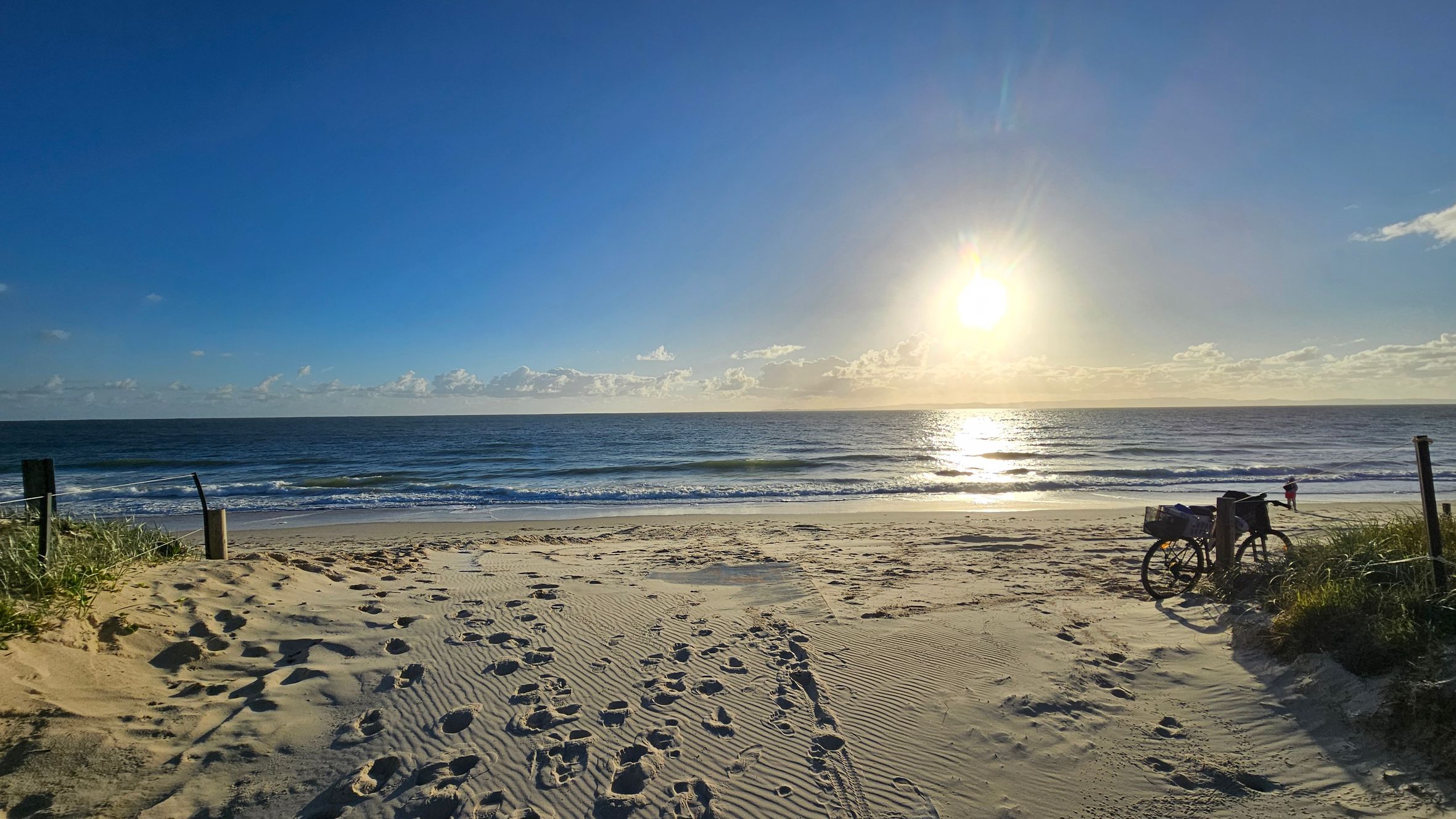 Sandy beach with footprints leading towards the ocean. The sun is low on the horizon, casting a bright reflection on the water. On the right, a bicycle is parked near some grass and a wooden post. A person stands near the water's edge. The sky is clear with a few clouds near the horizon.