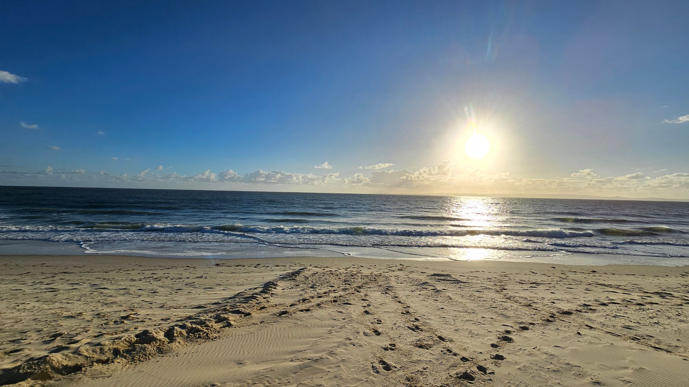 Sandy beach in the foreground with visible footprints and tyre tracks. The ocean stretches across the middle, with gentle waves approaching the shore. The sun is low on the horizon, casting a bright reflection on the water. A few clouds are scattered near the horizon, and the sky is clear and blue.