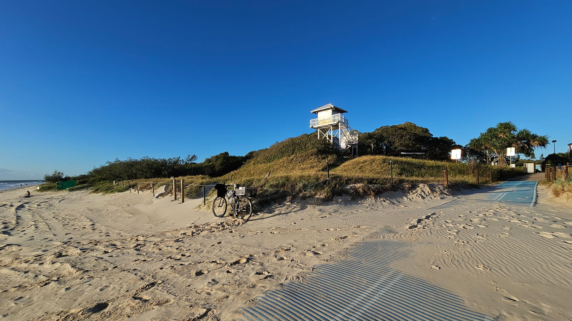 A sandy beach with a bicycle parked on the left near a path. The path leads to a grassy dune topped with a white lifeguard tower on the right. The area is surrounded by low shrubs and trees. The sand is marked with footprints and tyre tracks. A blue matting covers part of the sand, leading towards the dune. The sky is clear and blue.