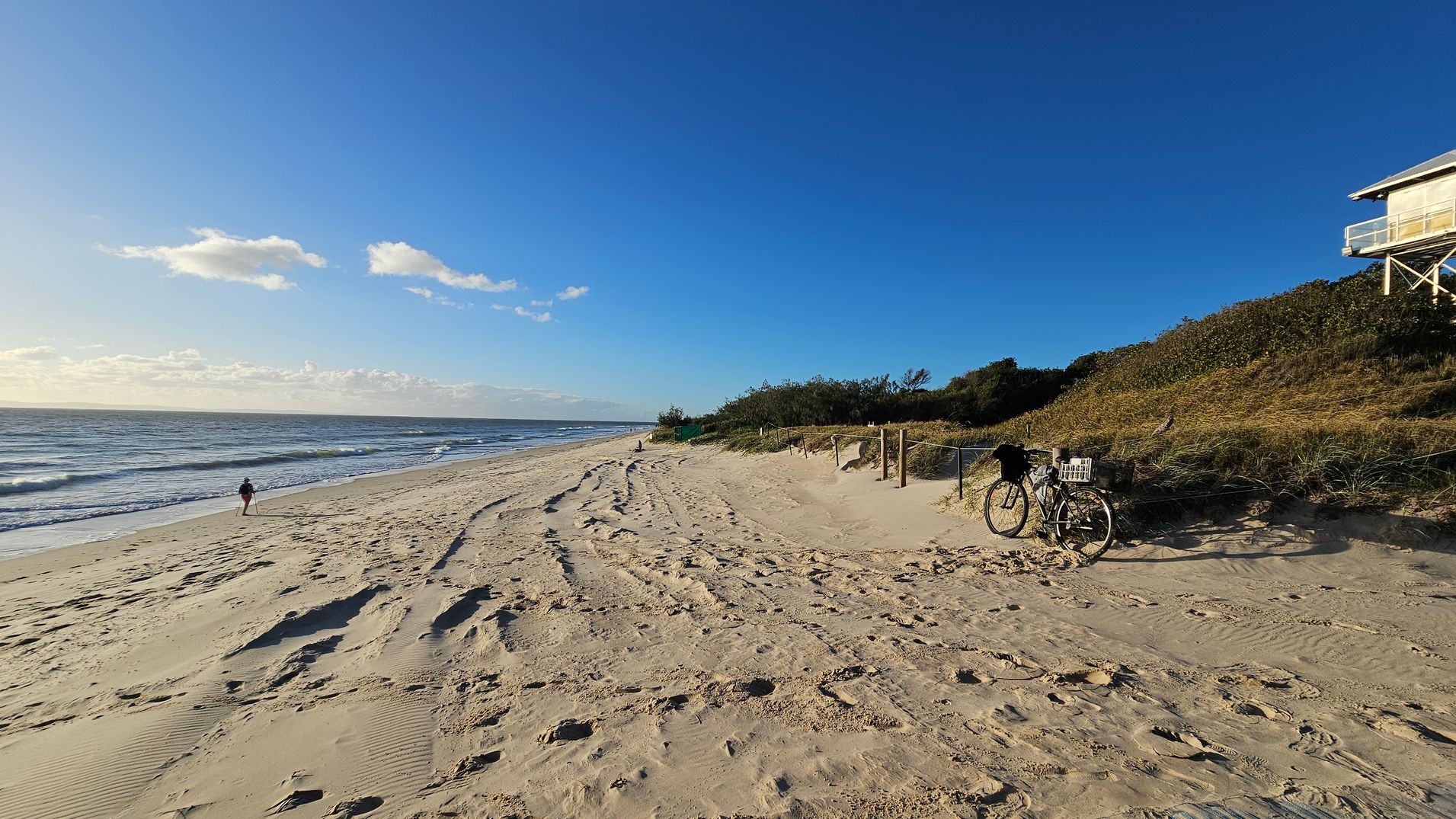 Sandy beach with gentle waves on the left. A person walks along the shoreline. On the right, a bicycle with a basket is parked near a grassy dune. A lifeguard tower is elevated on stilts above the dune. The sky is clear with a few clouds.