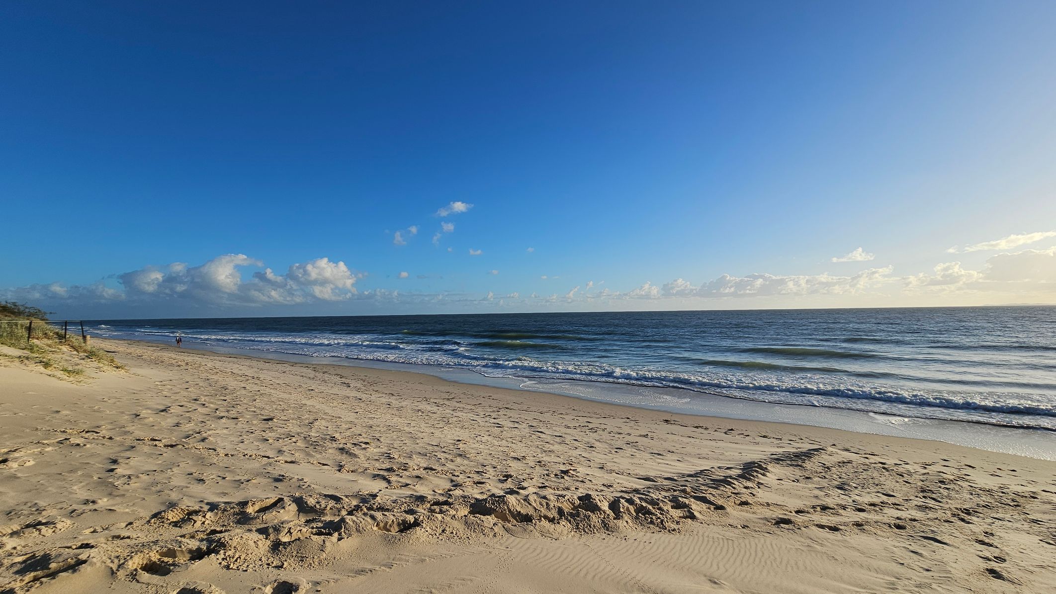 A sandy beach stretches across the foreground with footprints visible in the sand. On the left, there are small dunes with sparse vegetation and a fence. The ocean extends across the middle, with gentle waves lapping at the shore. A person is walking along the water's edge in the distance. The sky is clear with a few scattered clouds.