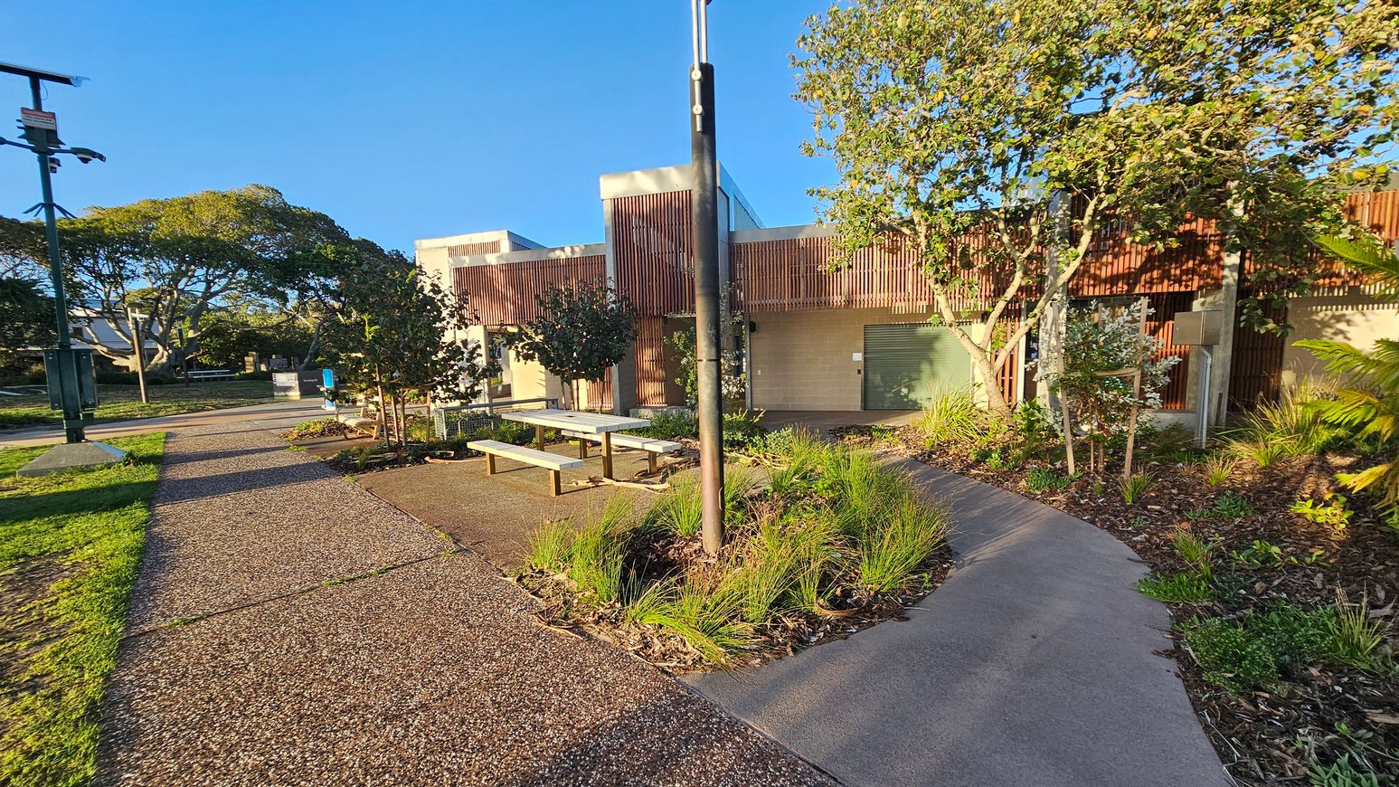A paved pathway curves through a landscaped area with grass and small plants. On the right, a modern building with wooden slats and a green door is partially visible. Trees and shrubs are planted around the building. In the centre, a tall pole stands near picnic tables. On the left, more trees and a grassy area are visible, with a lamp post and a small structure in the distance. The sky is clear and blue.