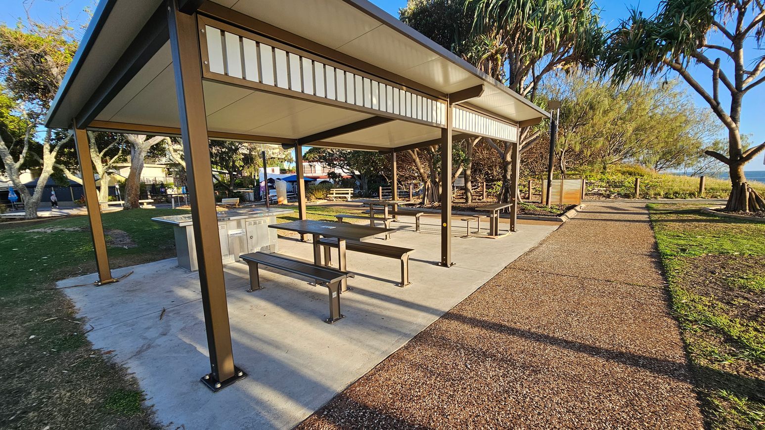A covered picnic area with metal benches and tables on a concrete floor. The structure has a flat roof supported by brown metal posts. To the right, a gravel path leads towards a grassy area with trees and shrubs. In the background, more trees and a glimpse of the ocean are visible. On the left, additional picnic tables are scattered on the grass, and a few people can be seen in the distance.