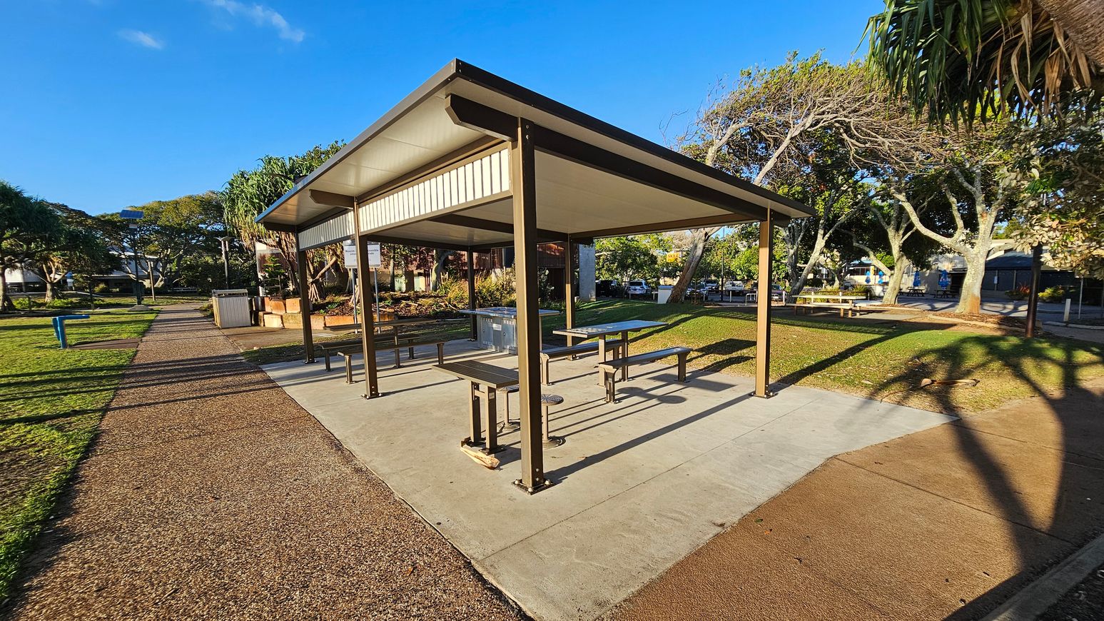 A park setting with a covered picnic area in the centre, featuring a metal roof supported by brown posts. Beneath the shelter are picnic tables and benches. The ground is a mix of concrete and gravel paths. To the left, a grassy area with a blue bin and trees. In the background, more trees and a few buildings are visible. Shadows of trees stretch across the ground, and the sky is clear and blue.