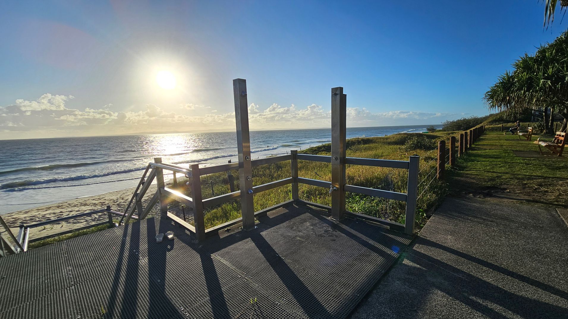 A wooden platform with railings in the foreground leads down to a sandy beach. The sun is low in the sky, casting long shadows and reflecting off the ocean. On the right, a grassy area with a path and wooden benches runs parallel to the beach, bordered by a fence. Vegetation, including trees, is visible along the path. The ocean stretches across the background, with gentle waves approaching the shore.