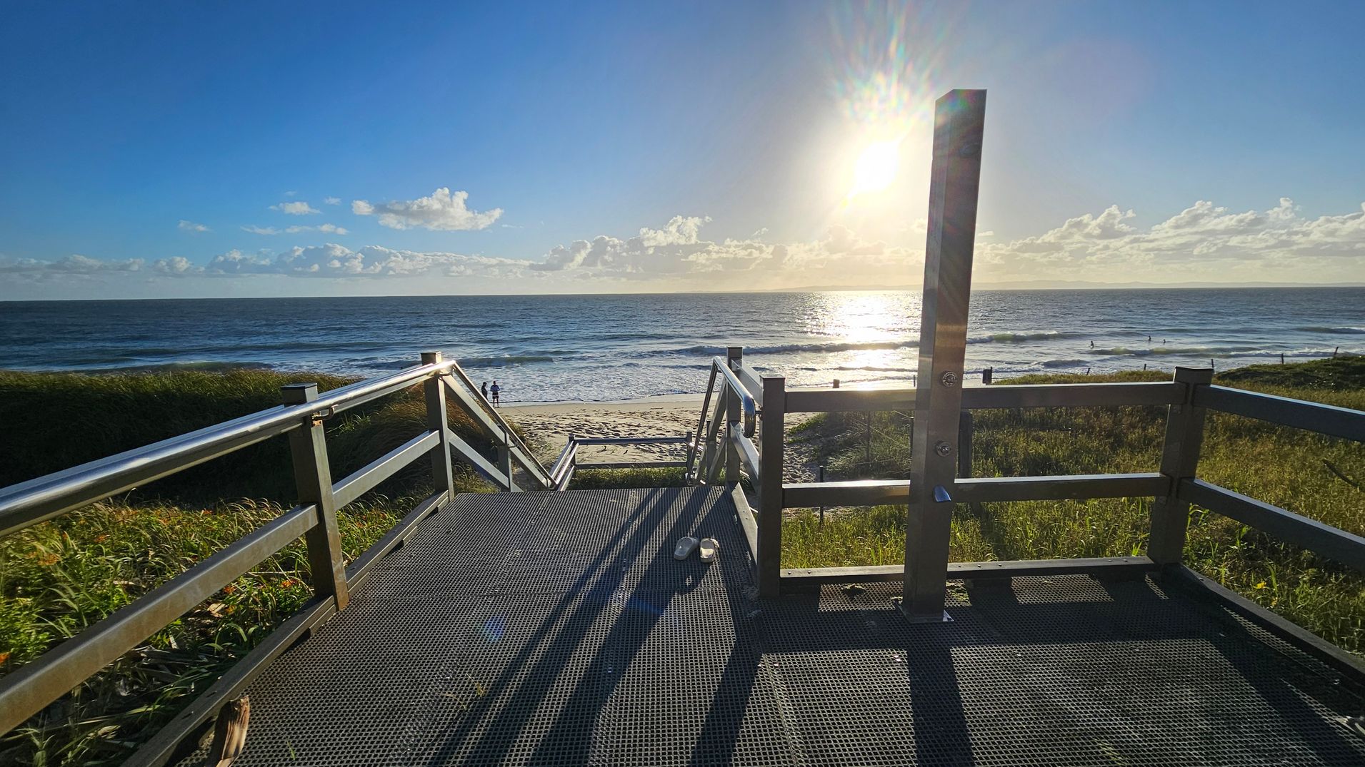A metal walkway with railings leads down to a sandy beach. The sun is low in the sky, casting long shadows on the walkway. On the left and right, grassy dunes border the path. In the distance, the ocean stretches out with gentle waves, and a few people are visible near the water's edge. A pair of shoes is placed on the walkway.