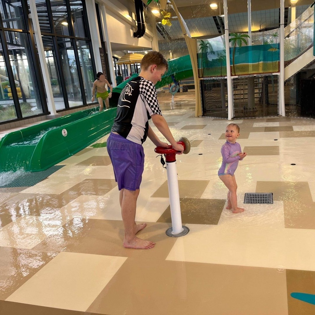 A boy helps a toddler at a water play area with a splash pad.